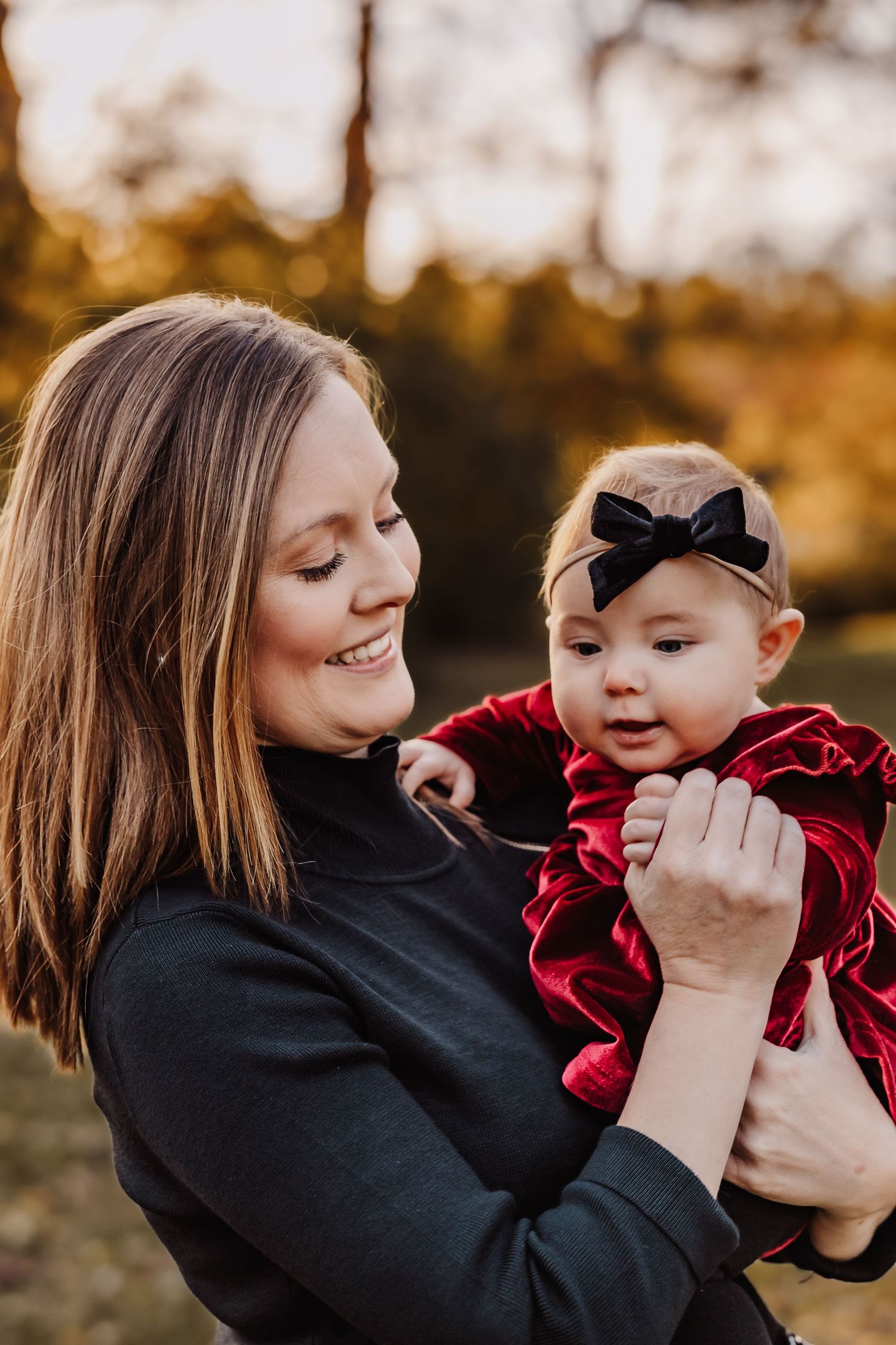 A tender moment captured between parent and baby in red dress with black bow against autumn background.