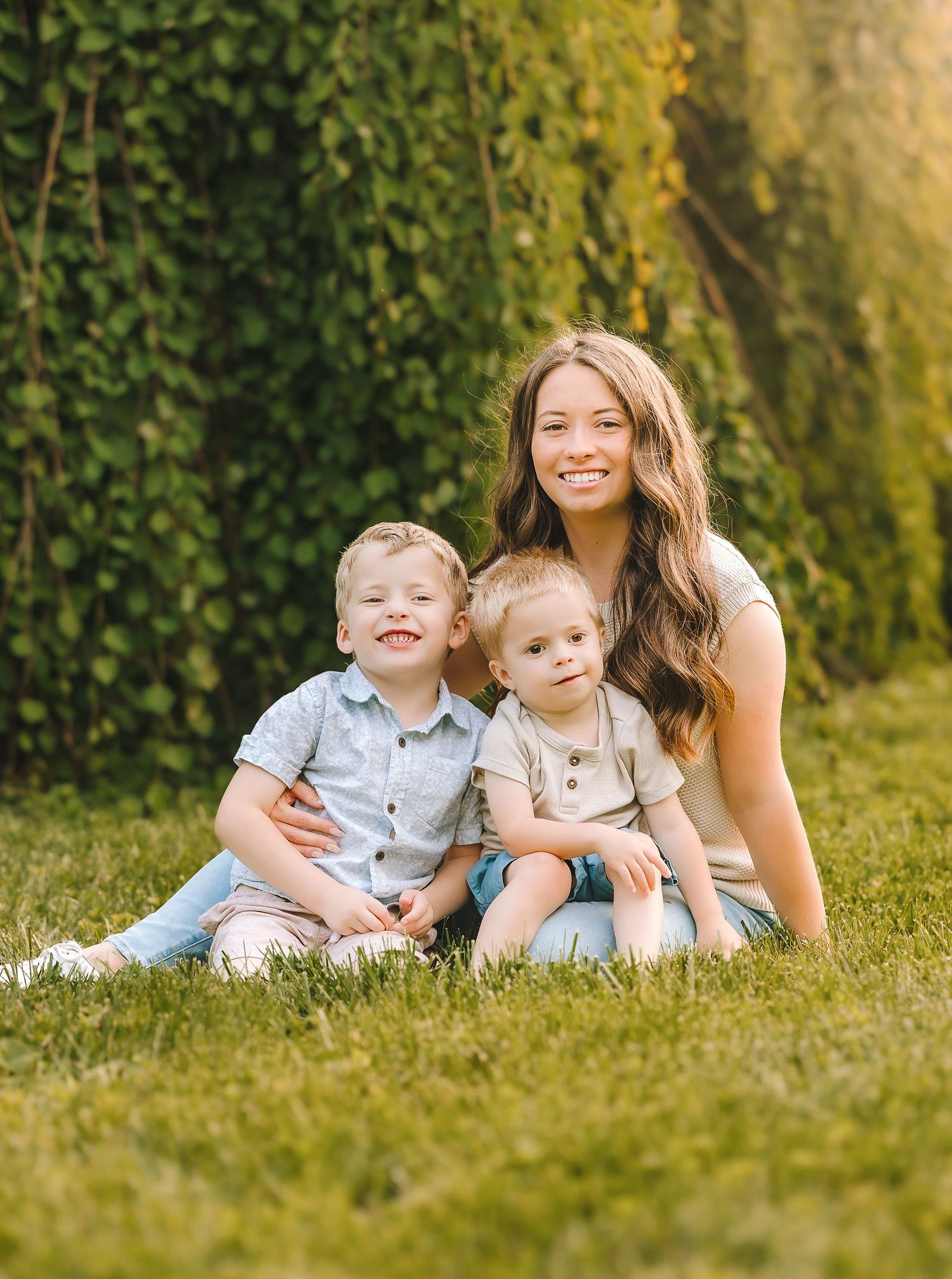 A family relaxes together on lush grass in front of a natural green backdrop during a sunny outdoor photo session.