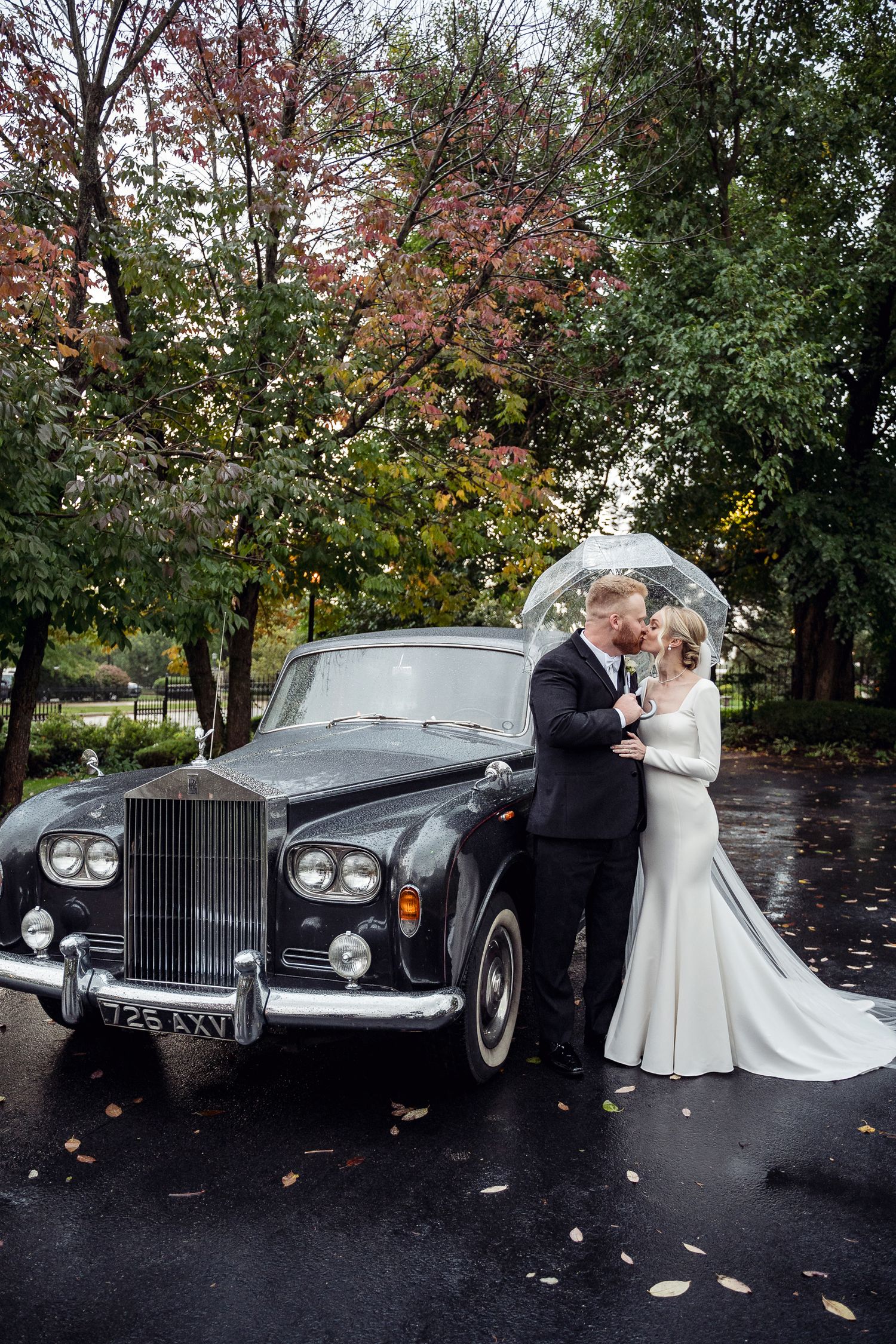 Bride and groom kissing under clear umbrella beside vintage black Rolls-Royce on rainy autumn day.