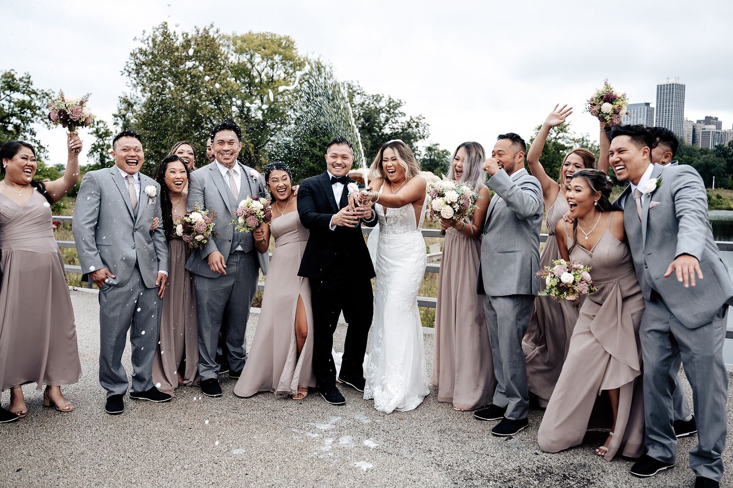 Wedding party in gray and blush attire celebrate together outdoors during a joyful group photo session.