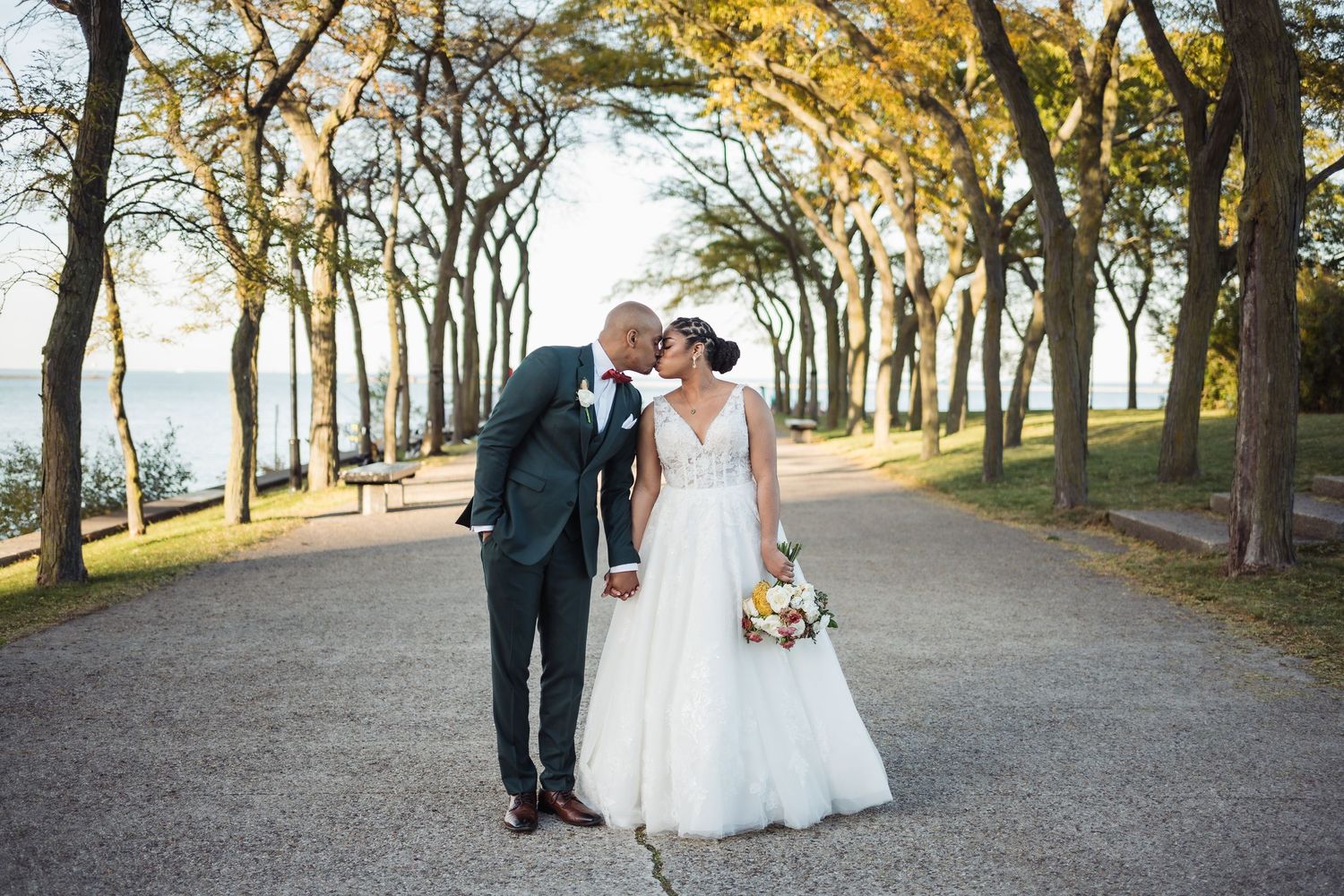 Bride and groom share a romantic kiss on a tree-lined path near a lake, bride holding a colorful bouquet.
