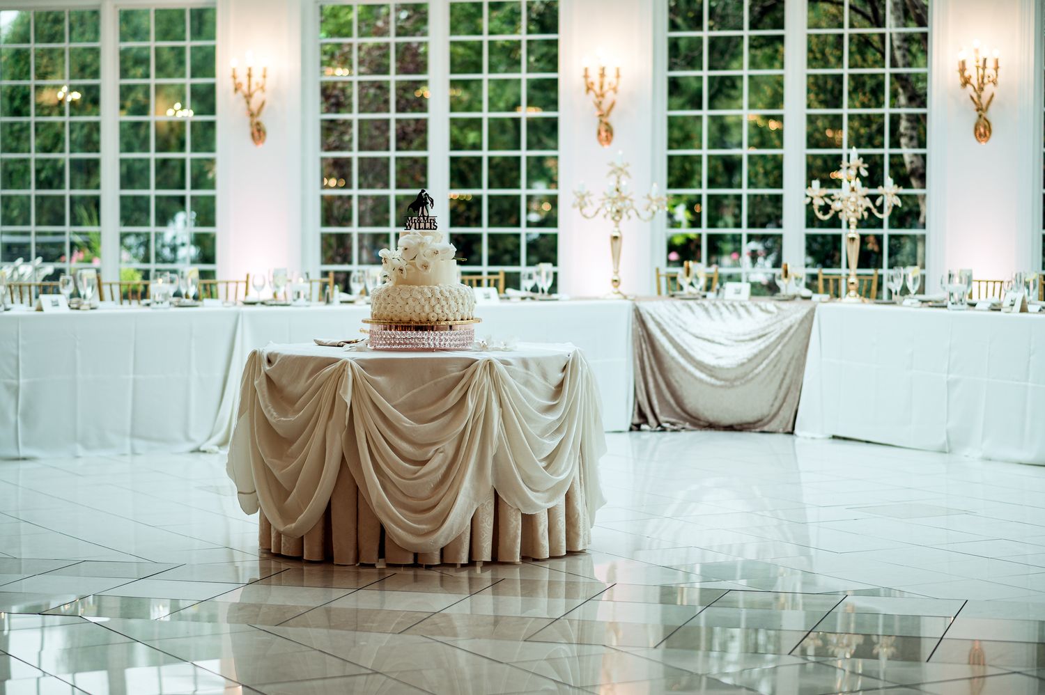 Elegant wedding reception with draped cake table, floral arrangements, and large windows in a bright ballroom.