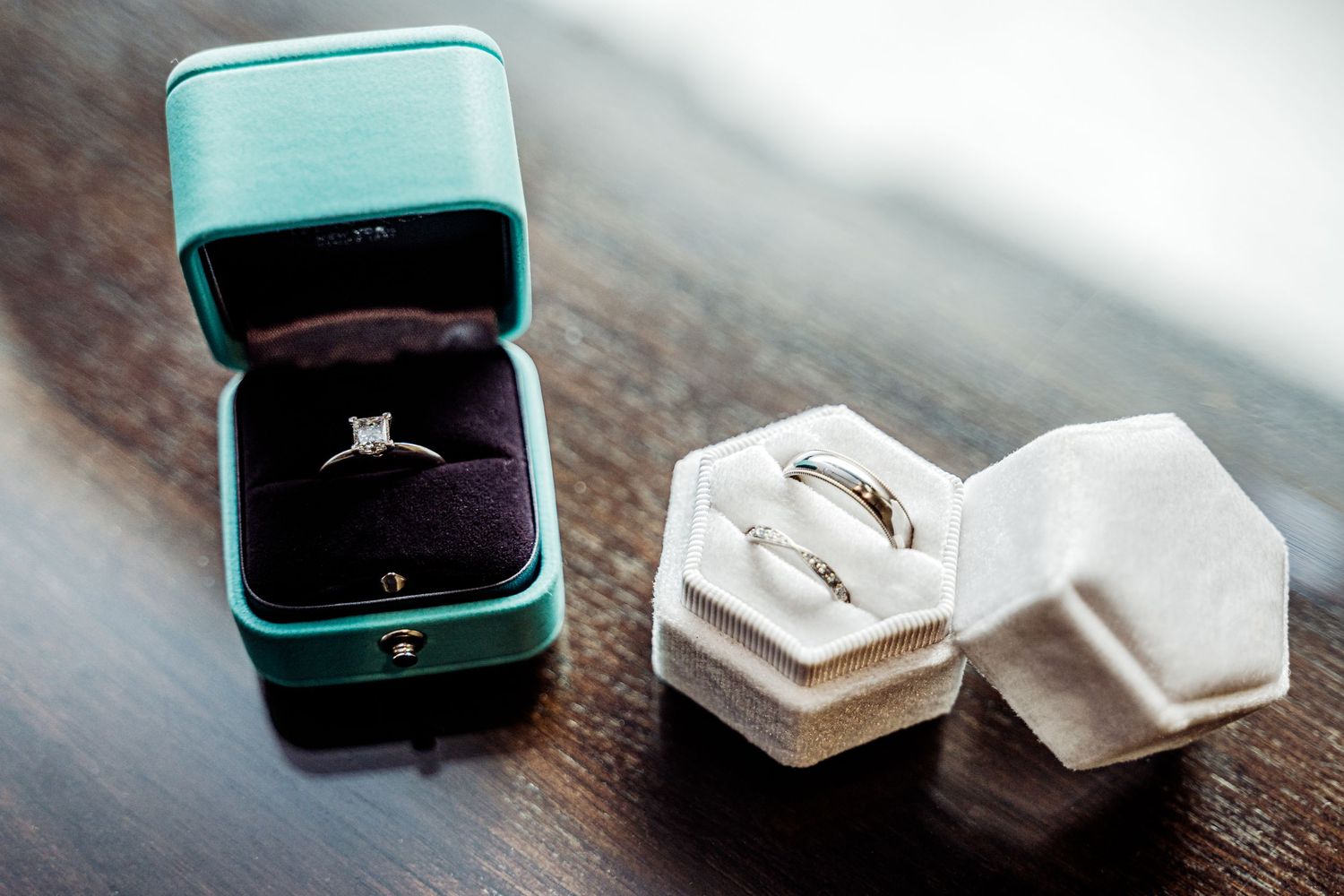Diamond engagement ring in teal box beside two wedding bands in white hexagon velvet box on wooden surface.