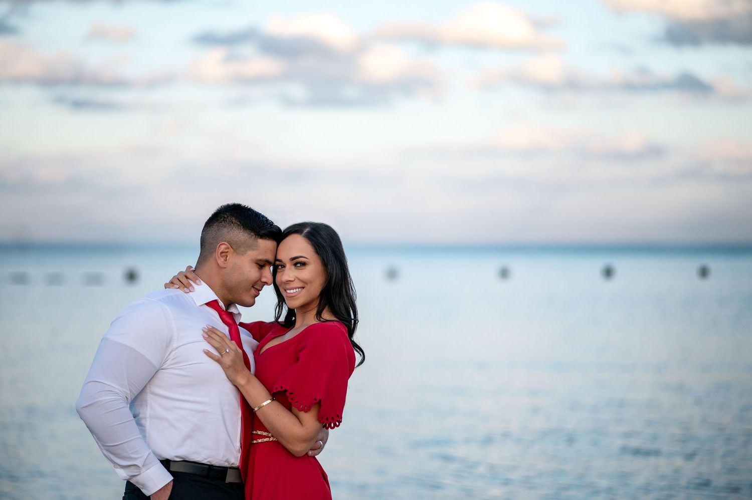 Couple embracing romantically by the ocean at sunset, woman in red dress smiling, man in white shirt, seaside backdrop.