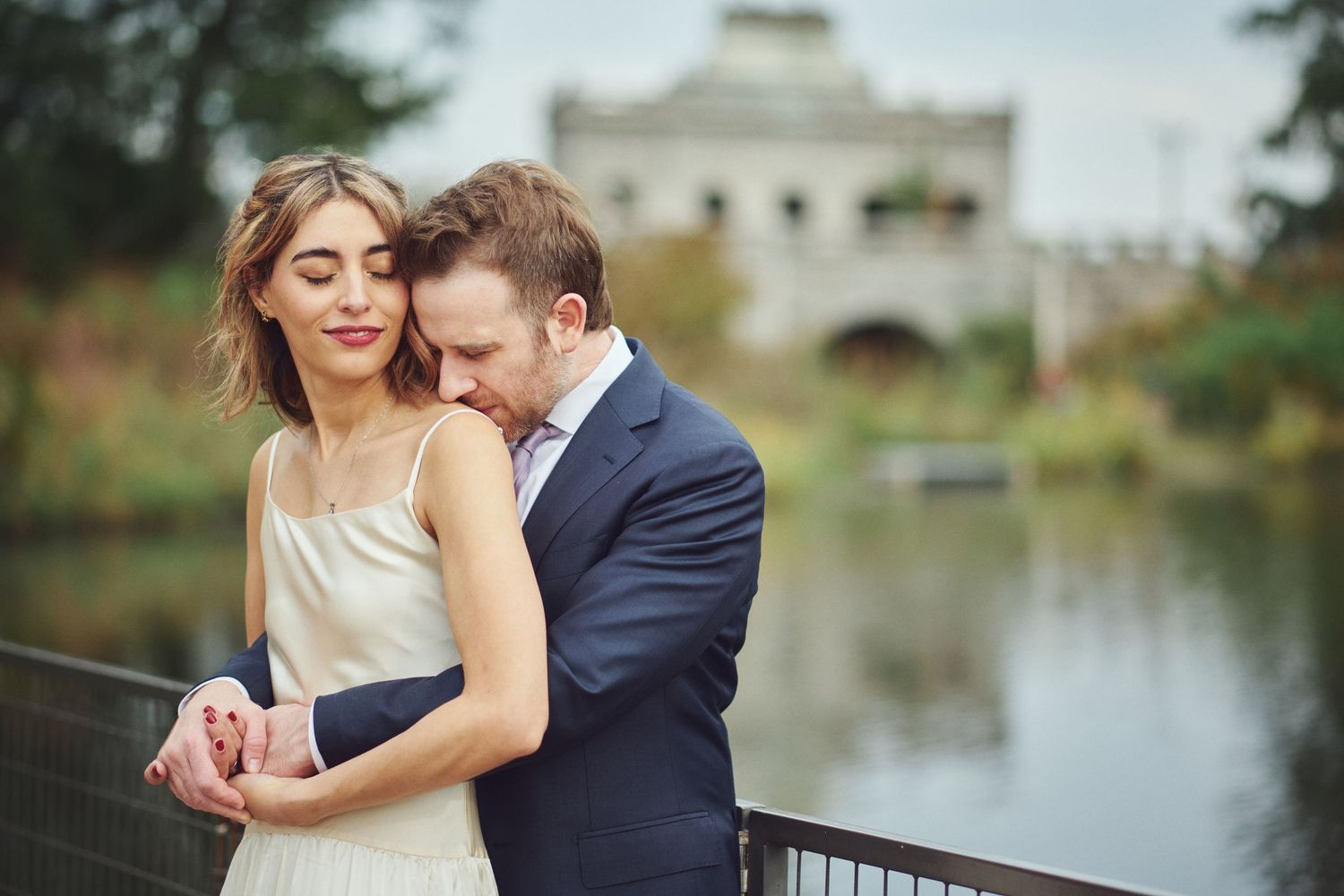 Romantic couple embracing on a bridge near a lake, man in navy suit nuzzling woman in elegant dress.