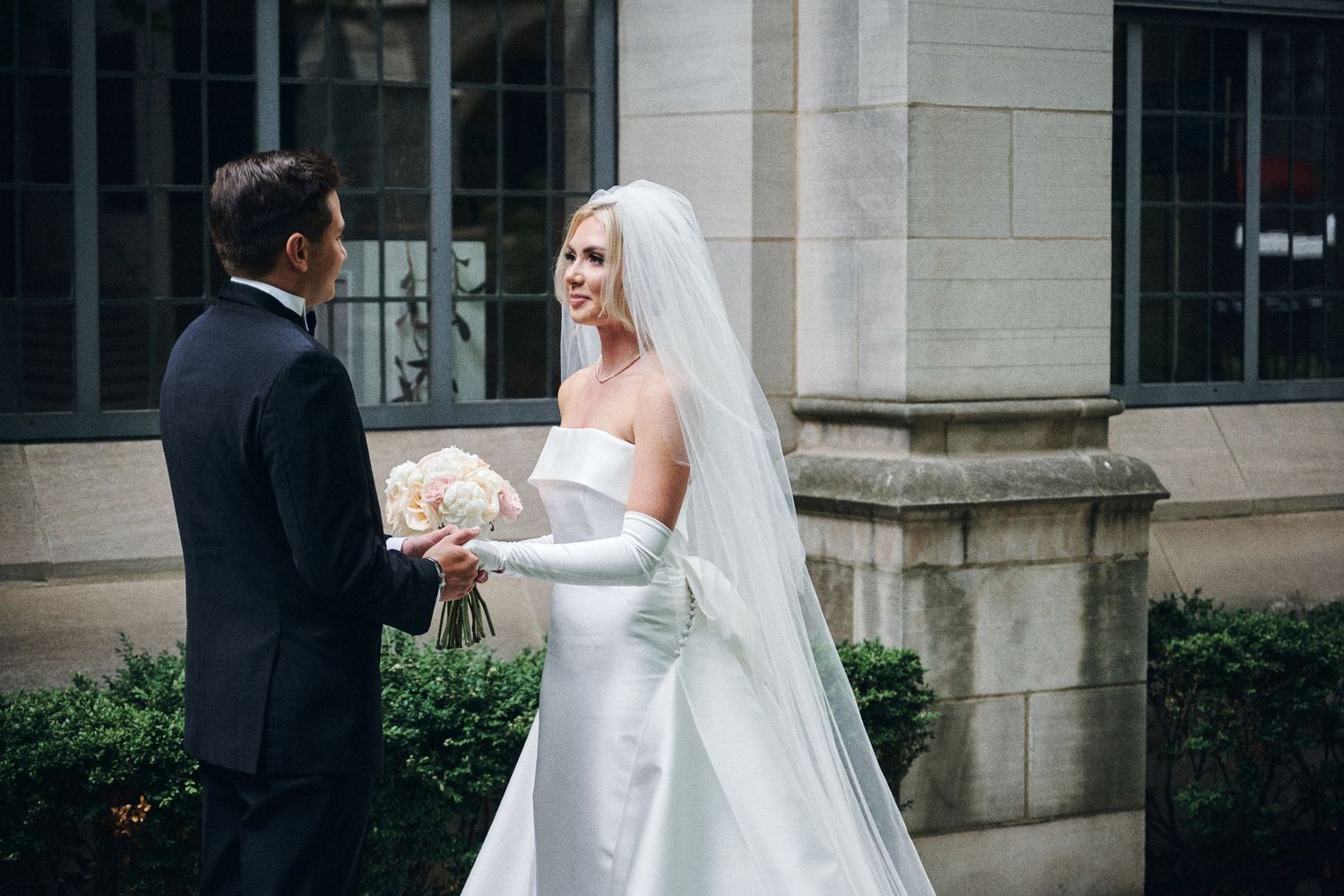 Bride in strapless white gown with veil holds bouquet while groom in tuxedo holds her hands outdoors.