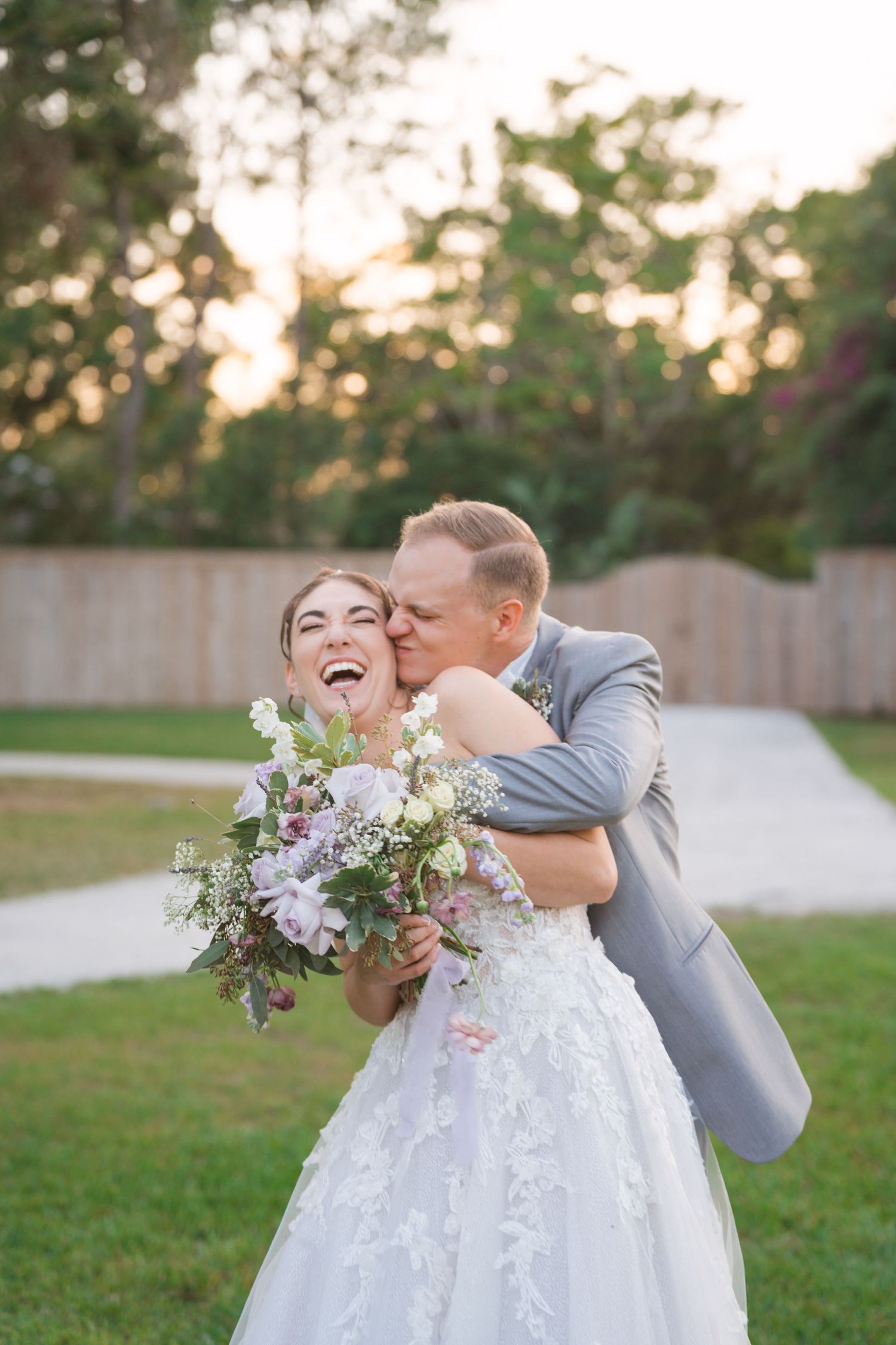 A romantic wedding photograph shows a couple embracing on a grassy lawn at sunset with a purple and white floral bouquet.