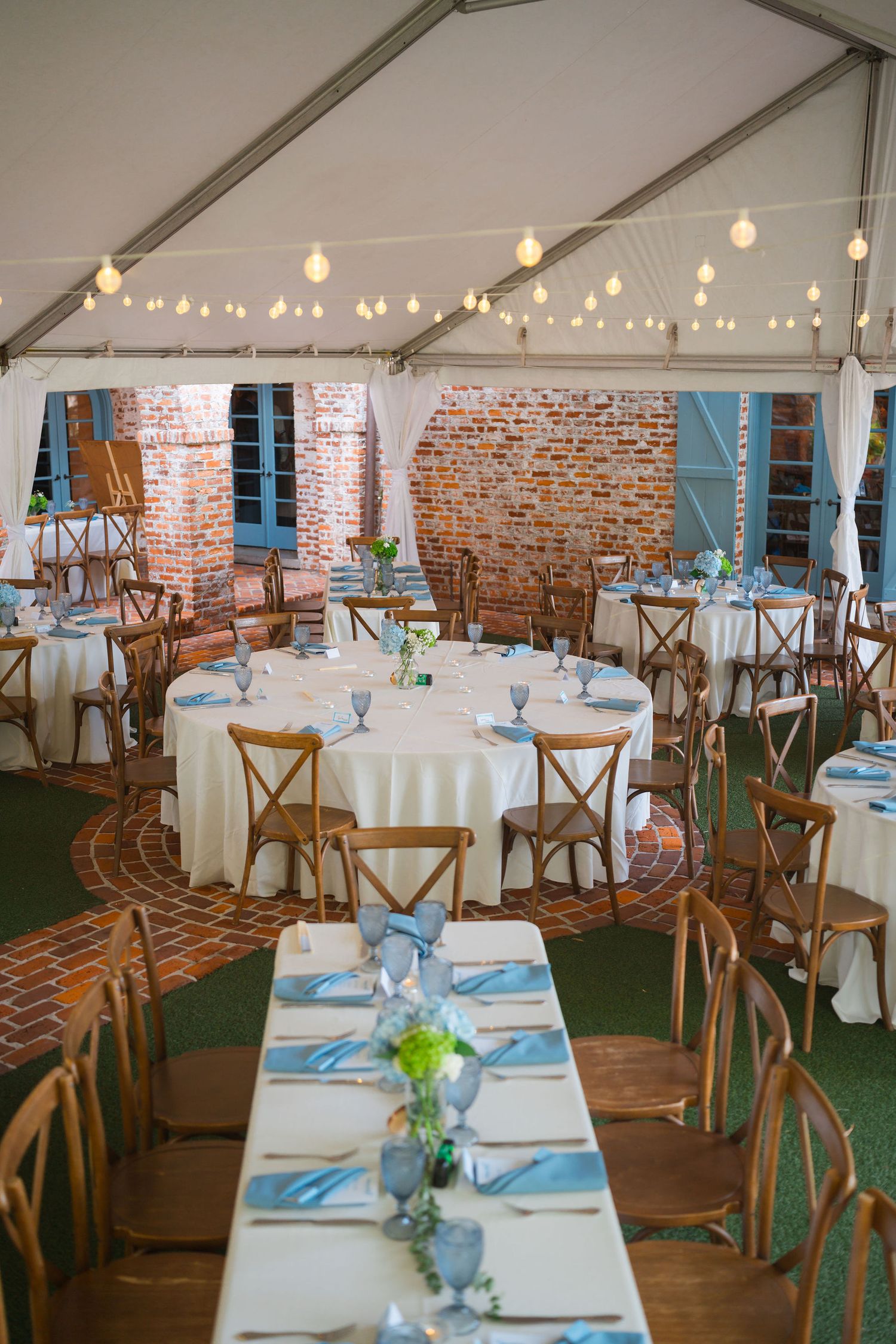Wedding reception setup with rustic wooden chairs, white tablecloths, and string lights under a tent against exposed brick walls.