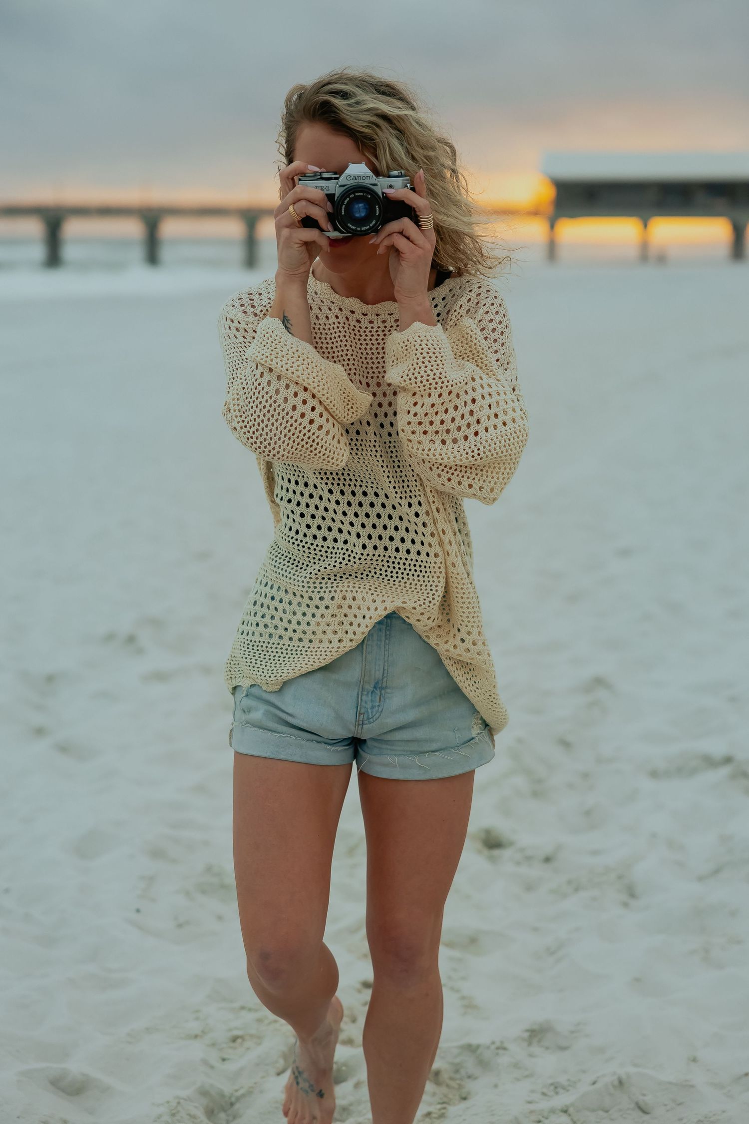 Person wearing cream crochet sweater and denim shorts poses on sandy beach at sunset with pier in background.