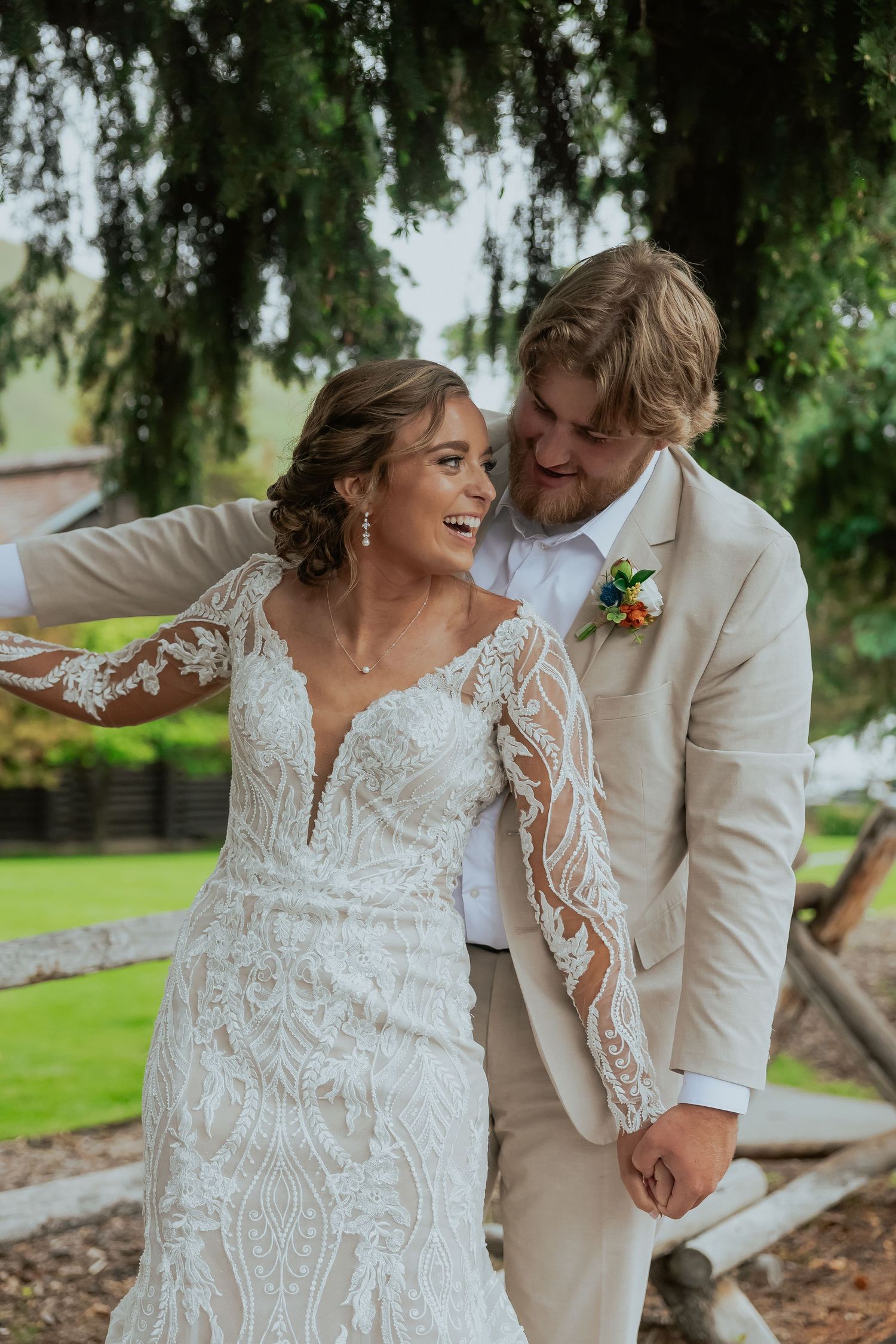 A couple shares a joyful moment under trees wearing wedding attire with lace details.