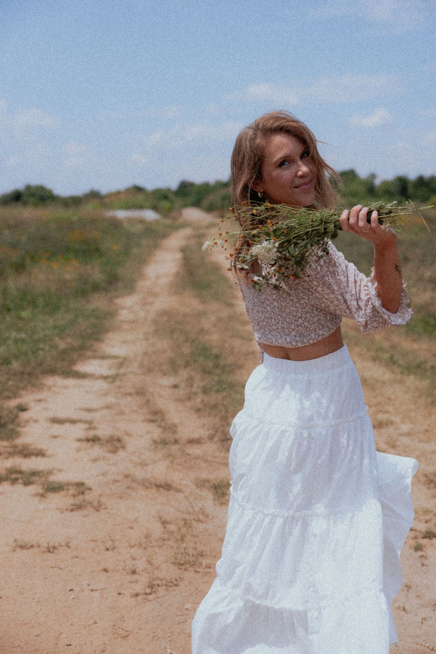 Someone in a white tiered maxi skirt and cropped top poses with wildflowers on a dirt path in a rural setting.