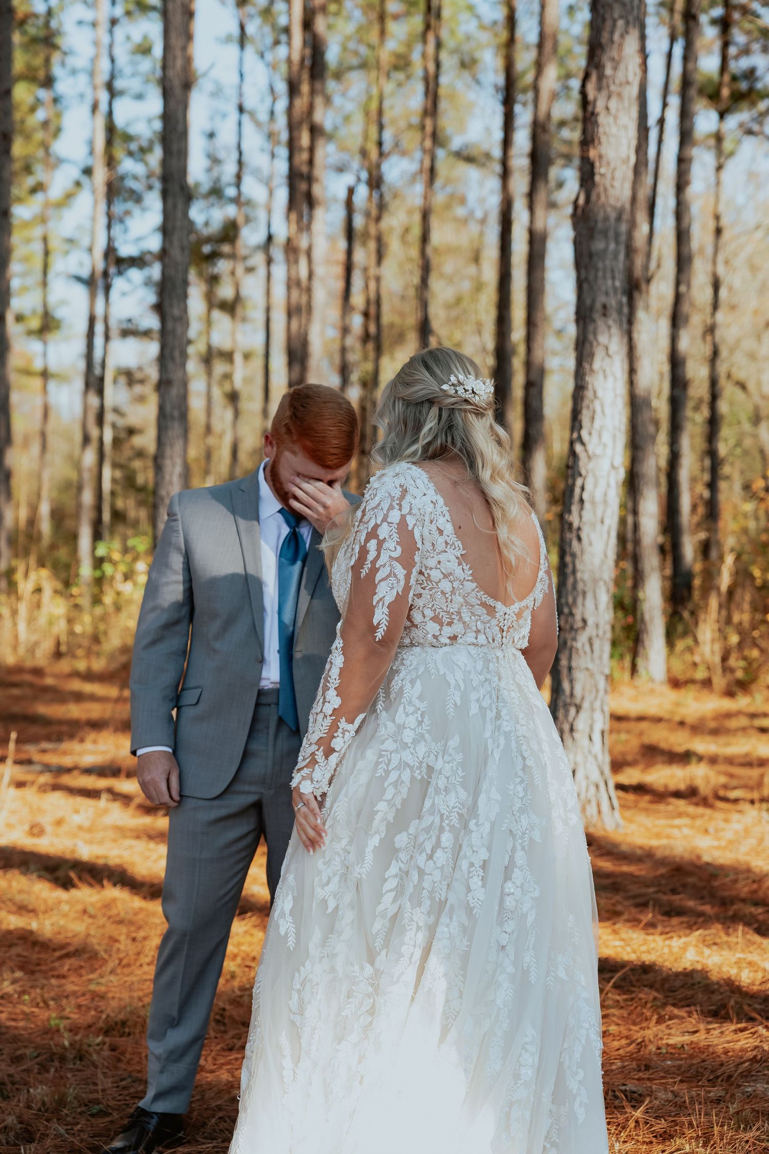 Couple in wedding attire walking through sun-lit pine forest.