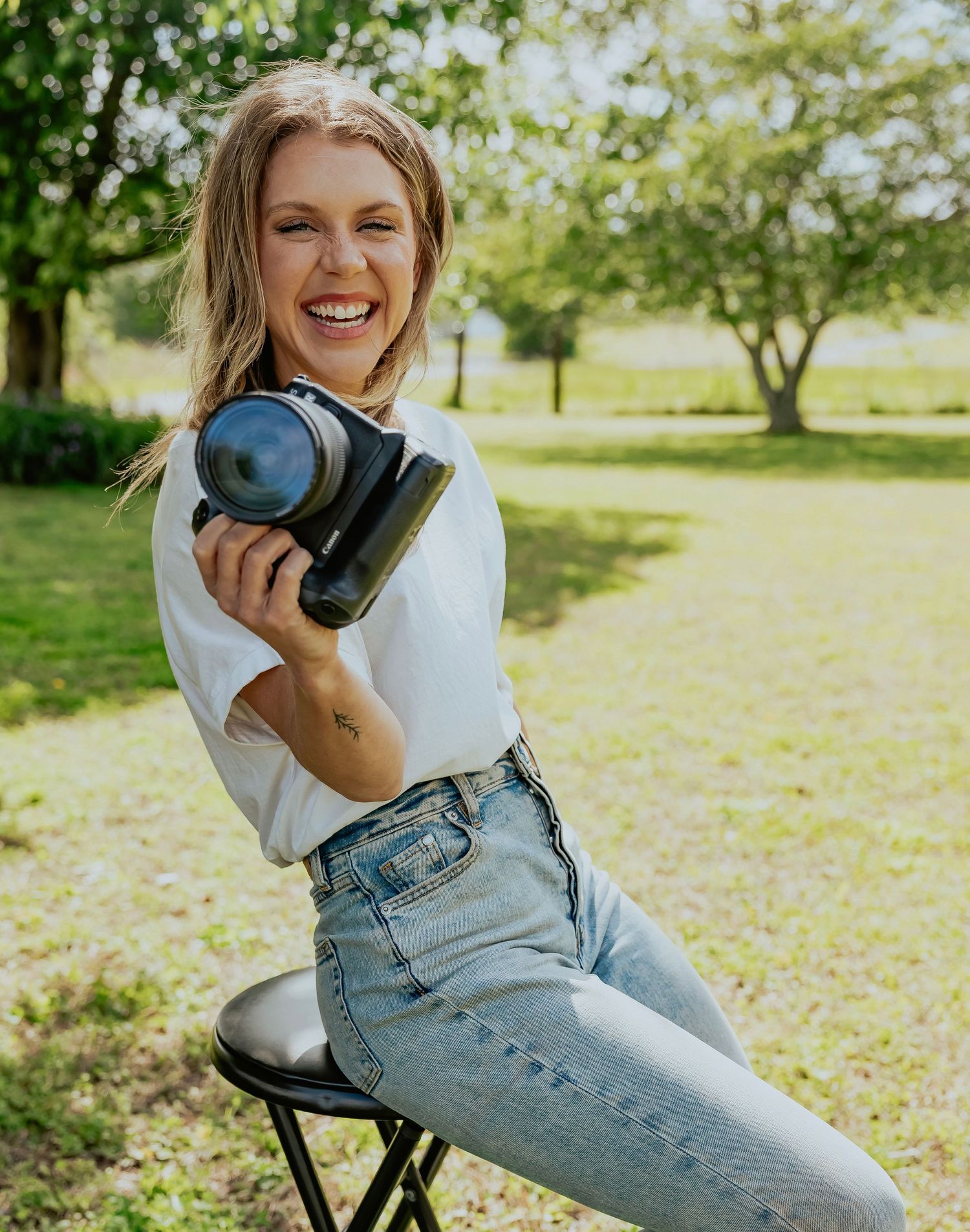 Person in white sweatshirt and light denim overalls poses playfully outdoors in a sunny park setting.