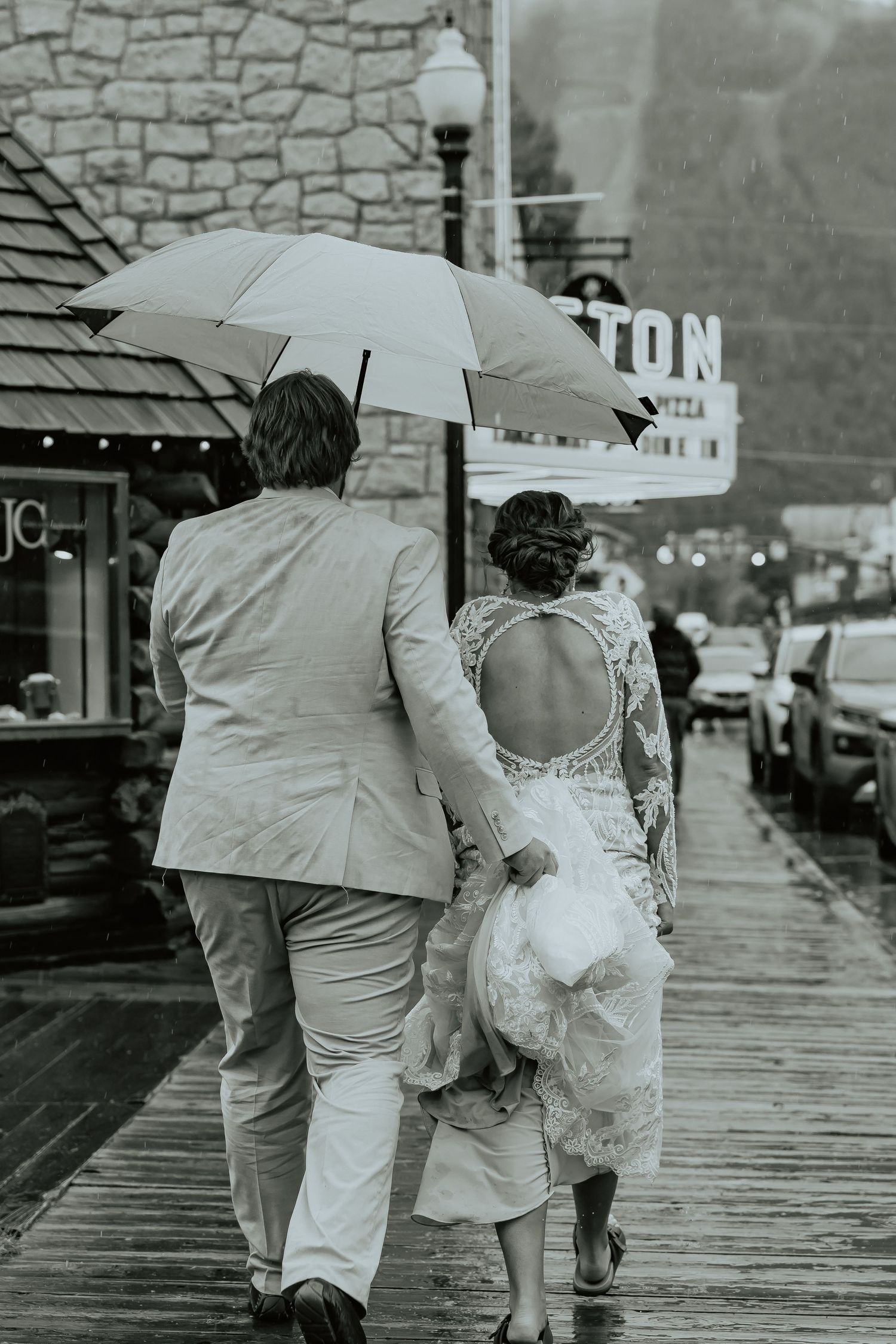 A couple walking together on a rainy boardwalk sharing an umbrella in a black and white photograph.