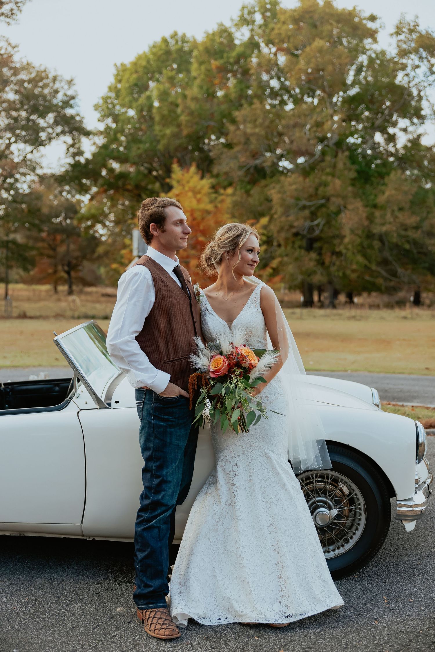 Bride and groom posing playfully next to vintage white car in autumn setting.