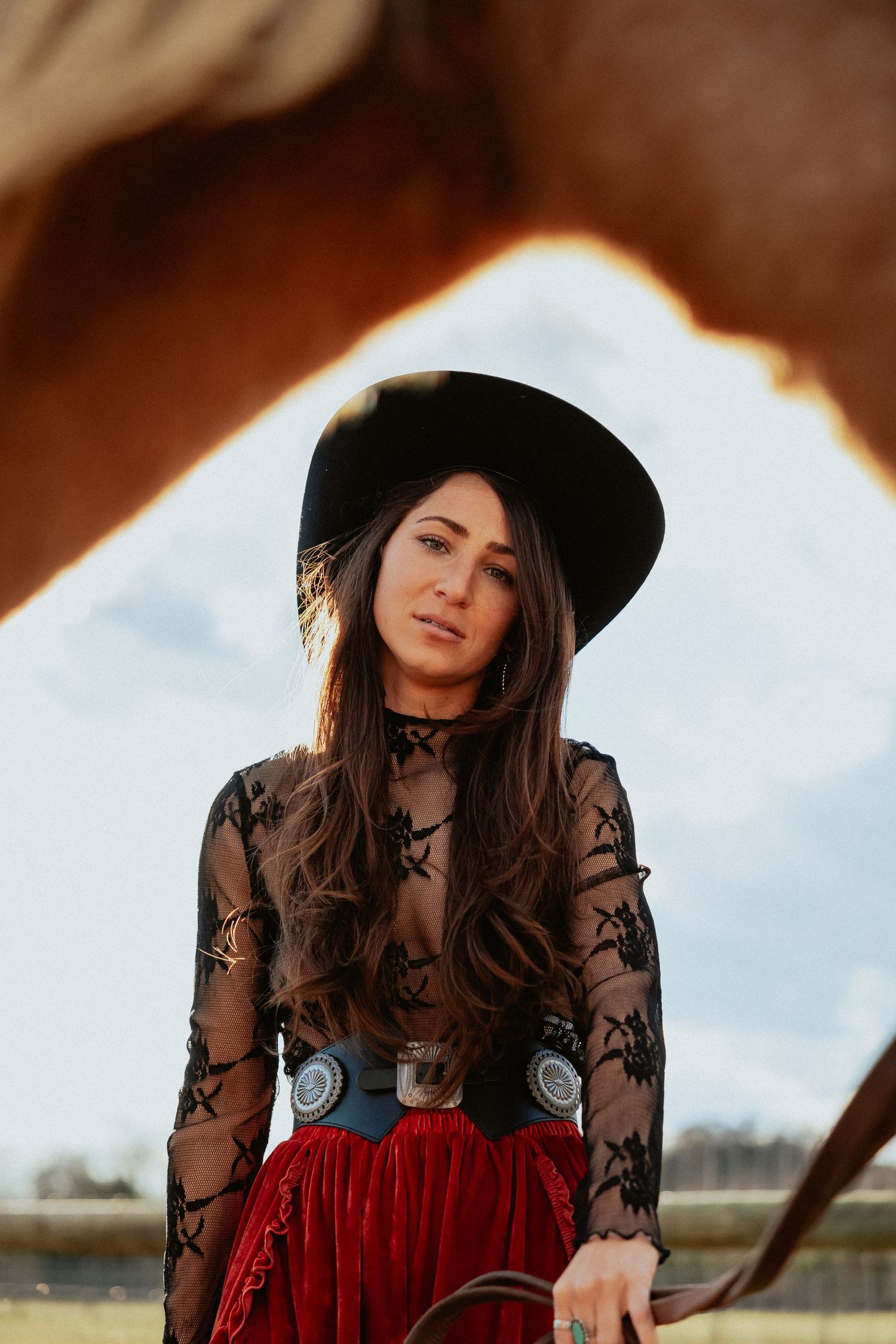 A woman in a black hat and lace top poses against a rustic outdoor setting with dramatic lighting.