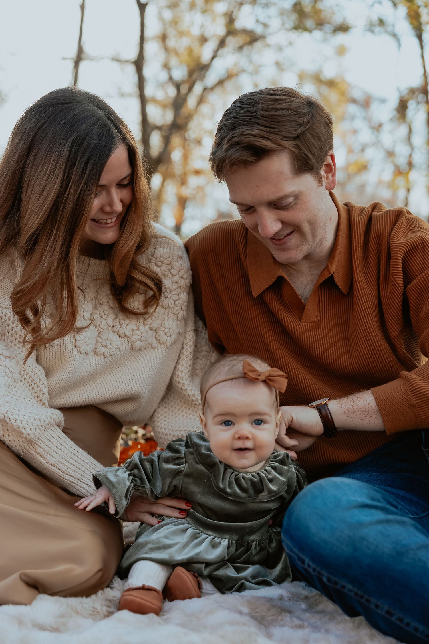 A couple in cozy sweaters sits with their baby wearing a green dress during an autumn outdoor photo session.