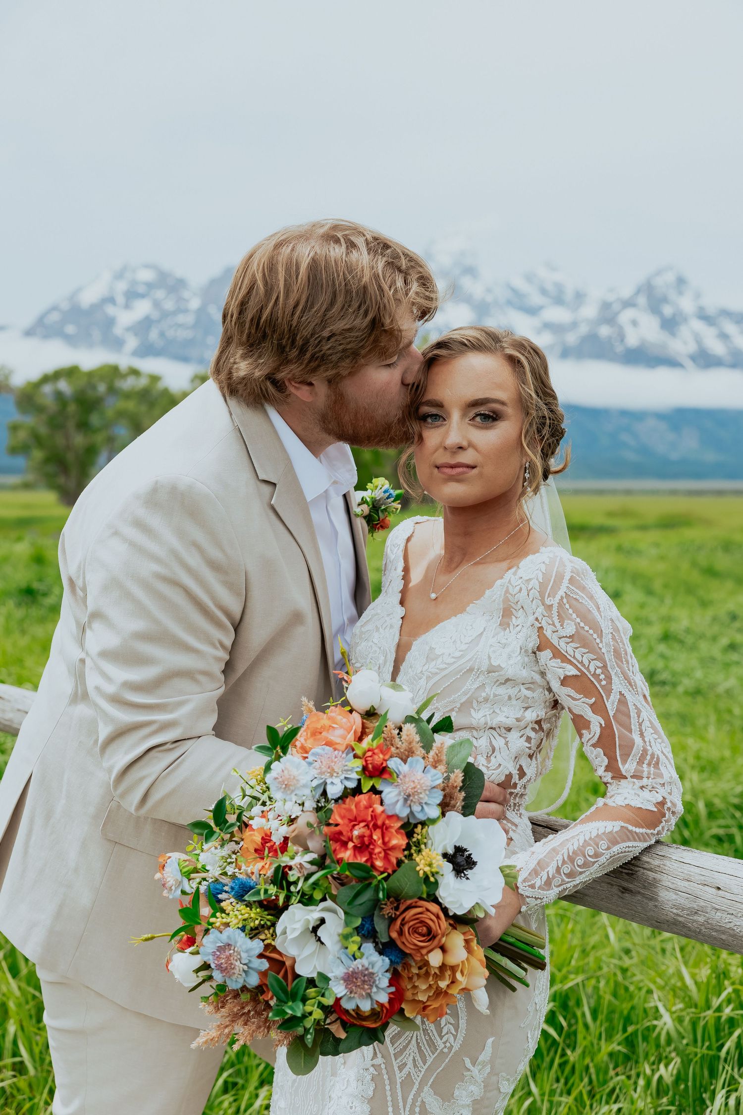 Couple embraces in meadow with mountain backdrop and vibrant orange and white floral bouquet.