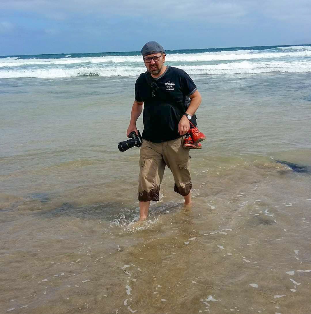Photographer crouching in shallow ocean waves with camera equipment on a sandy beach.