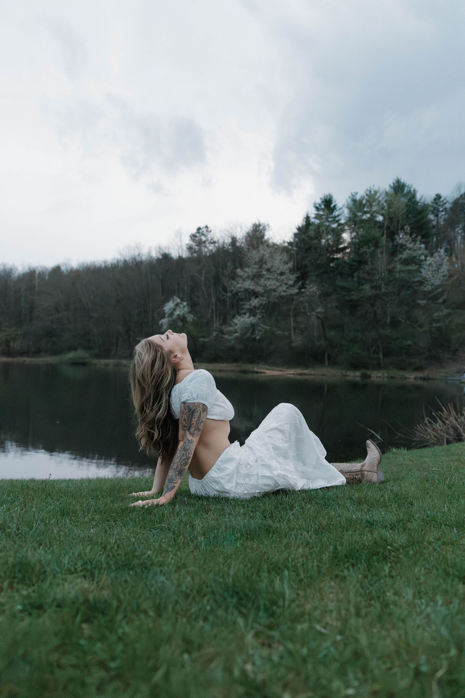 Someone in a white dress reclines on grassy lakeside with evergreen trees and cloudy skies reflecting in still water.