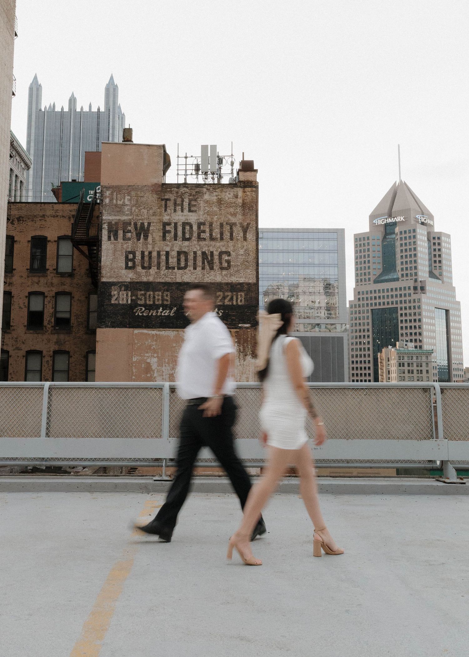 Two blurred figures walk across an urban rooftop with Pittsburgh skyscrapers and historic buildings in the background.