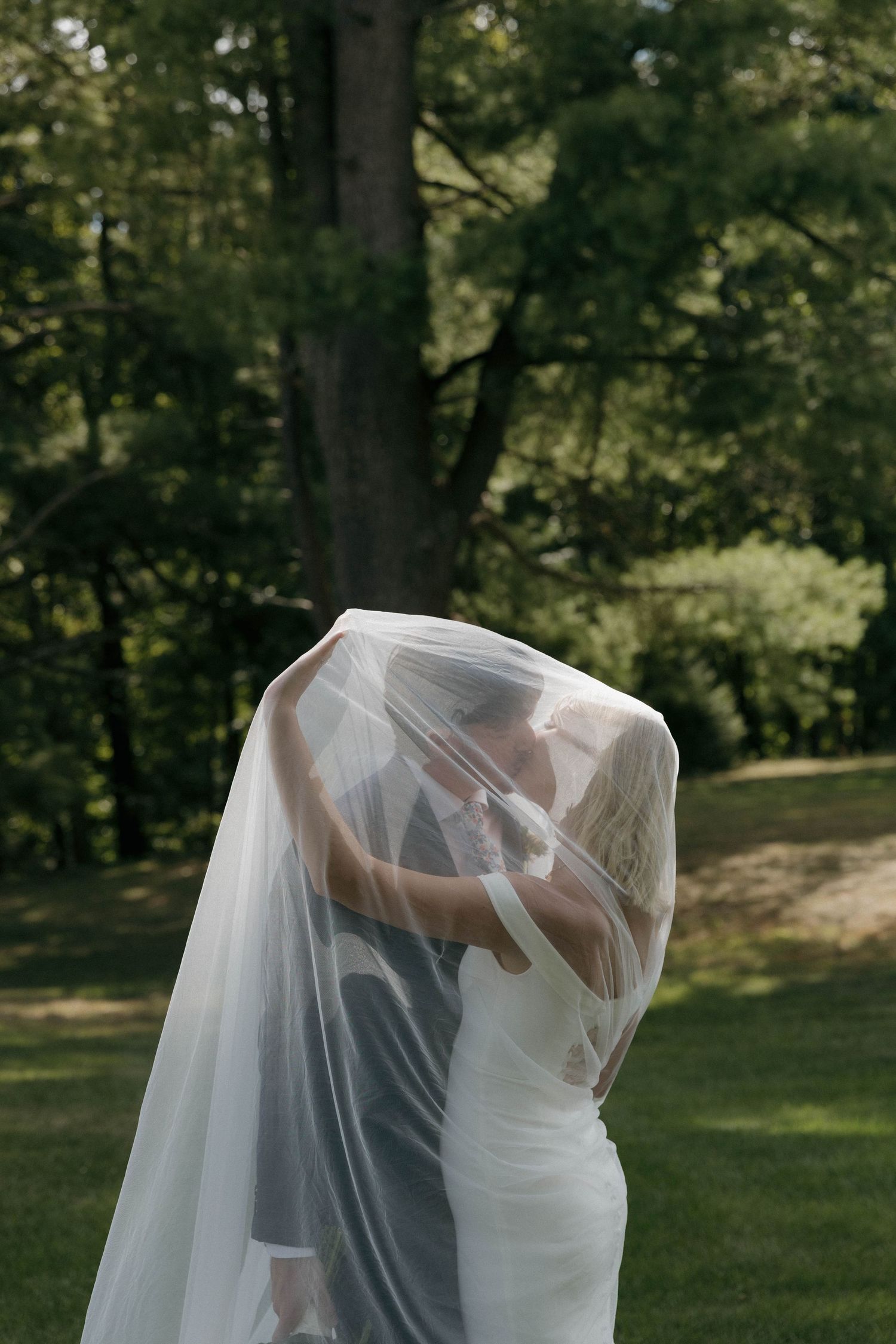 Romantic wedding couple embraces under flowing veil in lush garden setting during outdoor photo session.