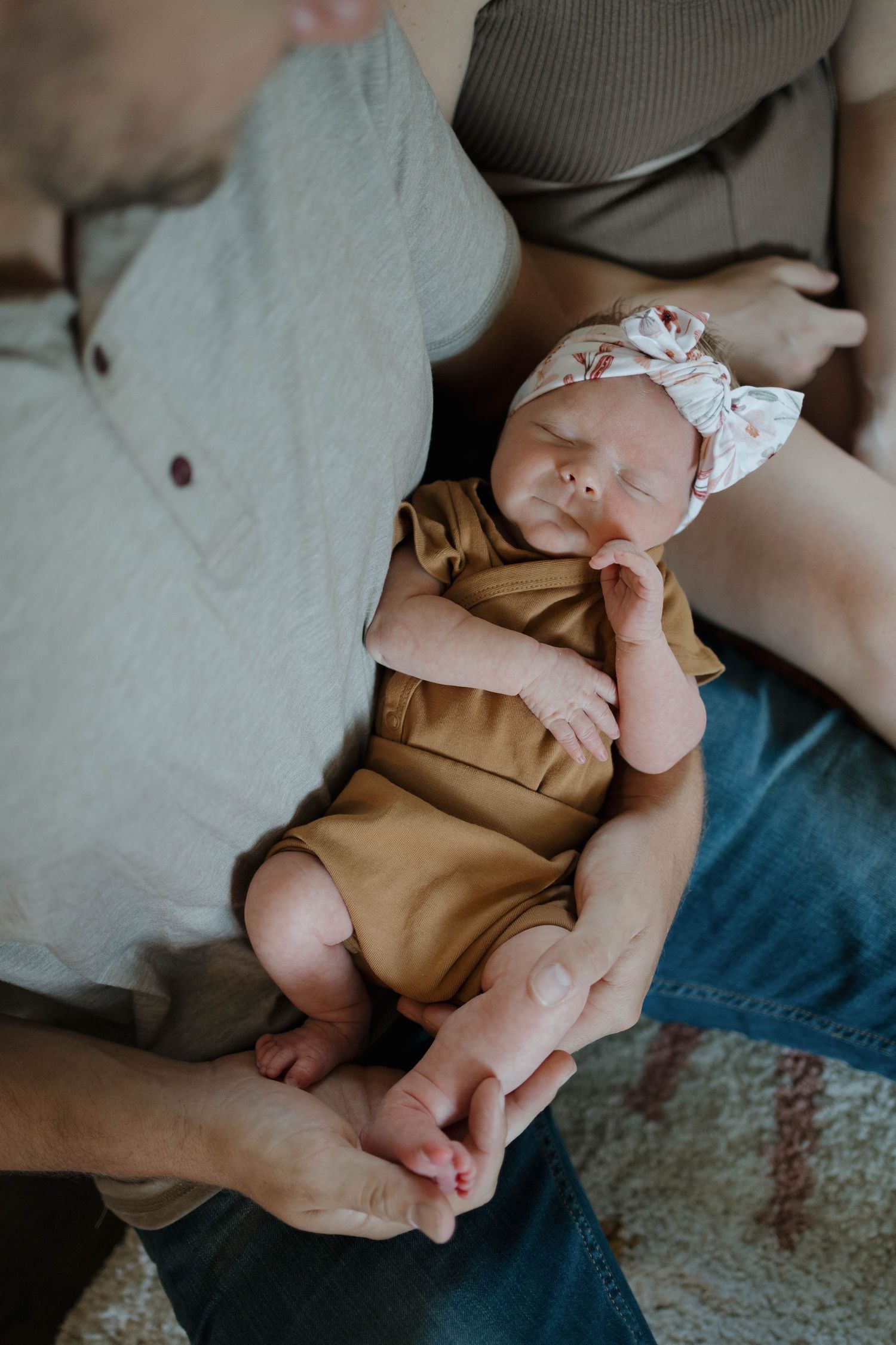 A sleeping baby wearing a brown romper and white bow headband rests comfortably on someone's lap.