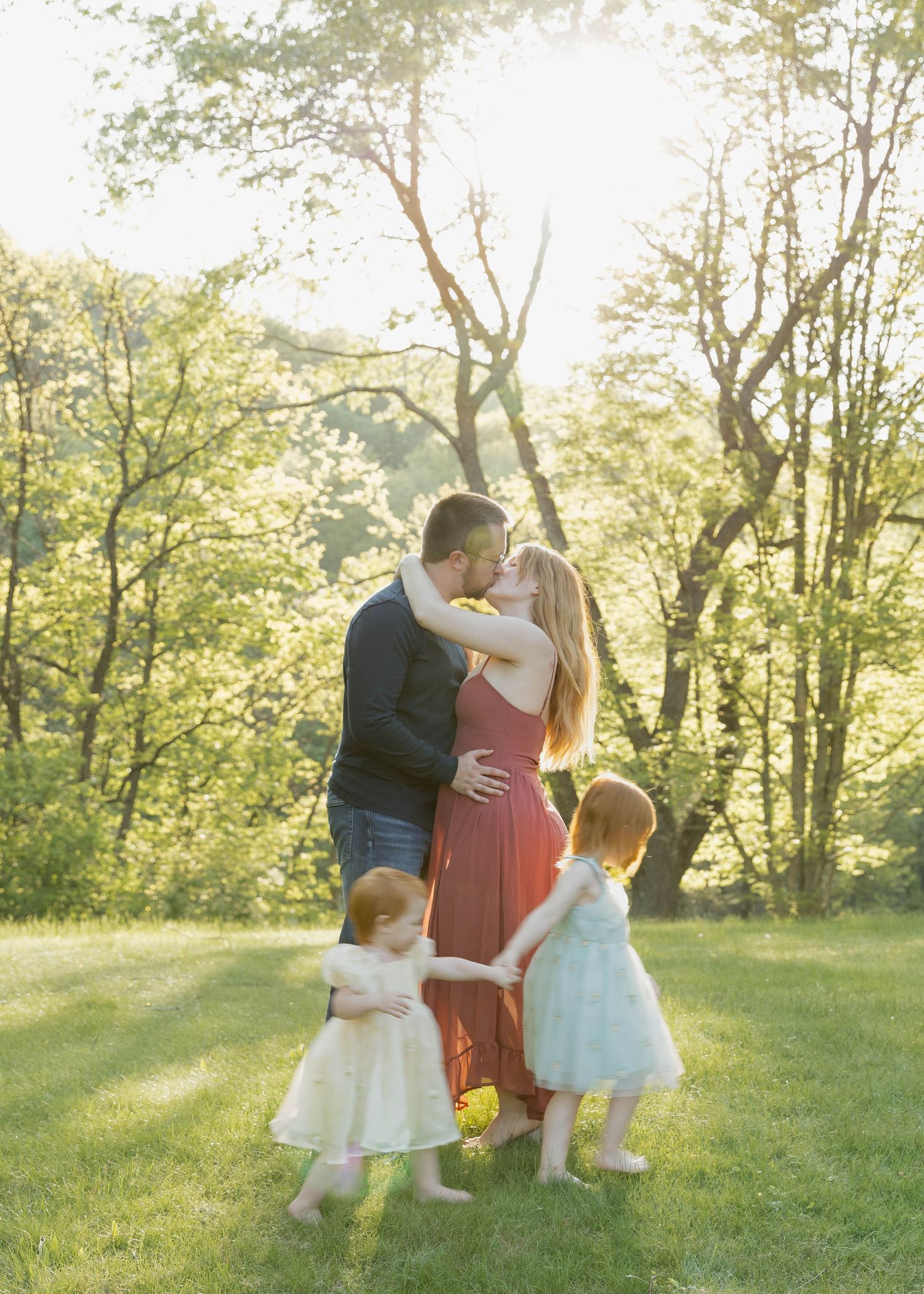 A loving couple embraces in a sunlit forest clearing while two small children in white dresses play around them.