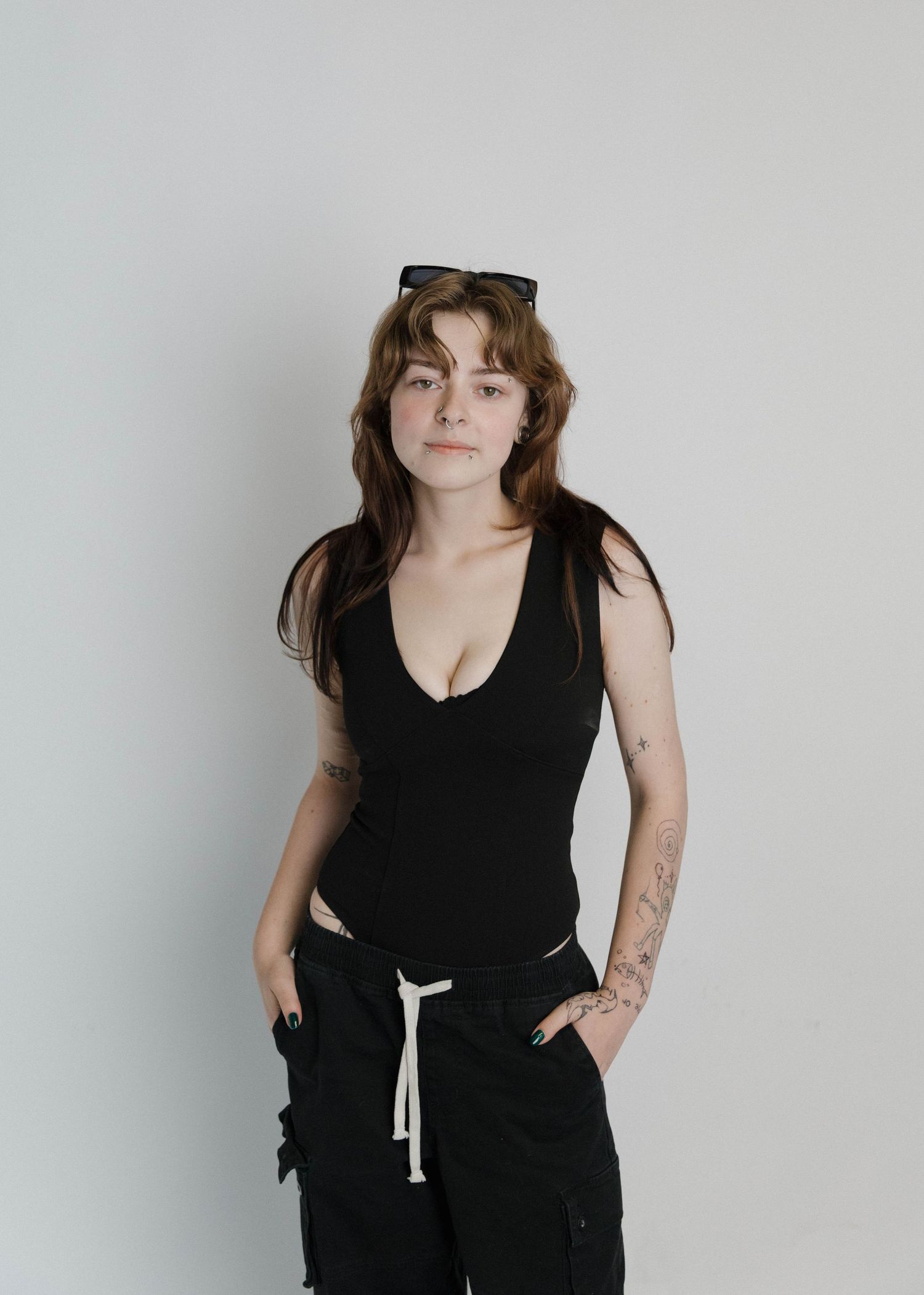 Person wearing a black sleeveless top and dark pants with sunglasses on head poses against plain wall in studio setting.