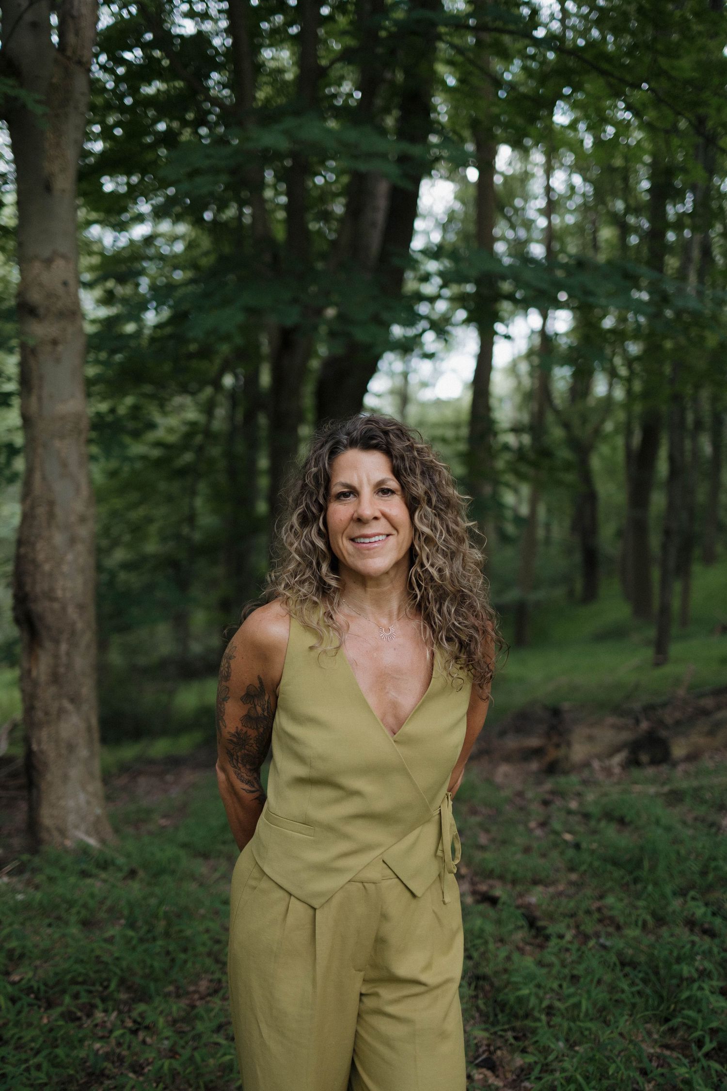 A person in an olive green jumpsuit poses outdoors in a lush forest setting with tall trees in the background.