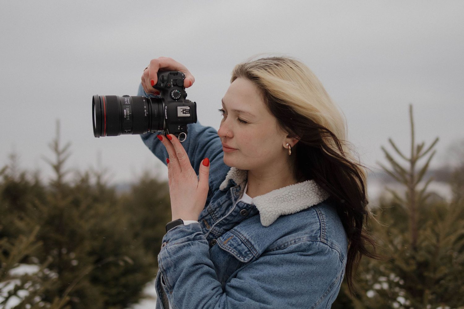 Person wearing denim jacket using camera outdoors with evergreen trees in background.