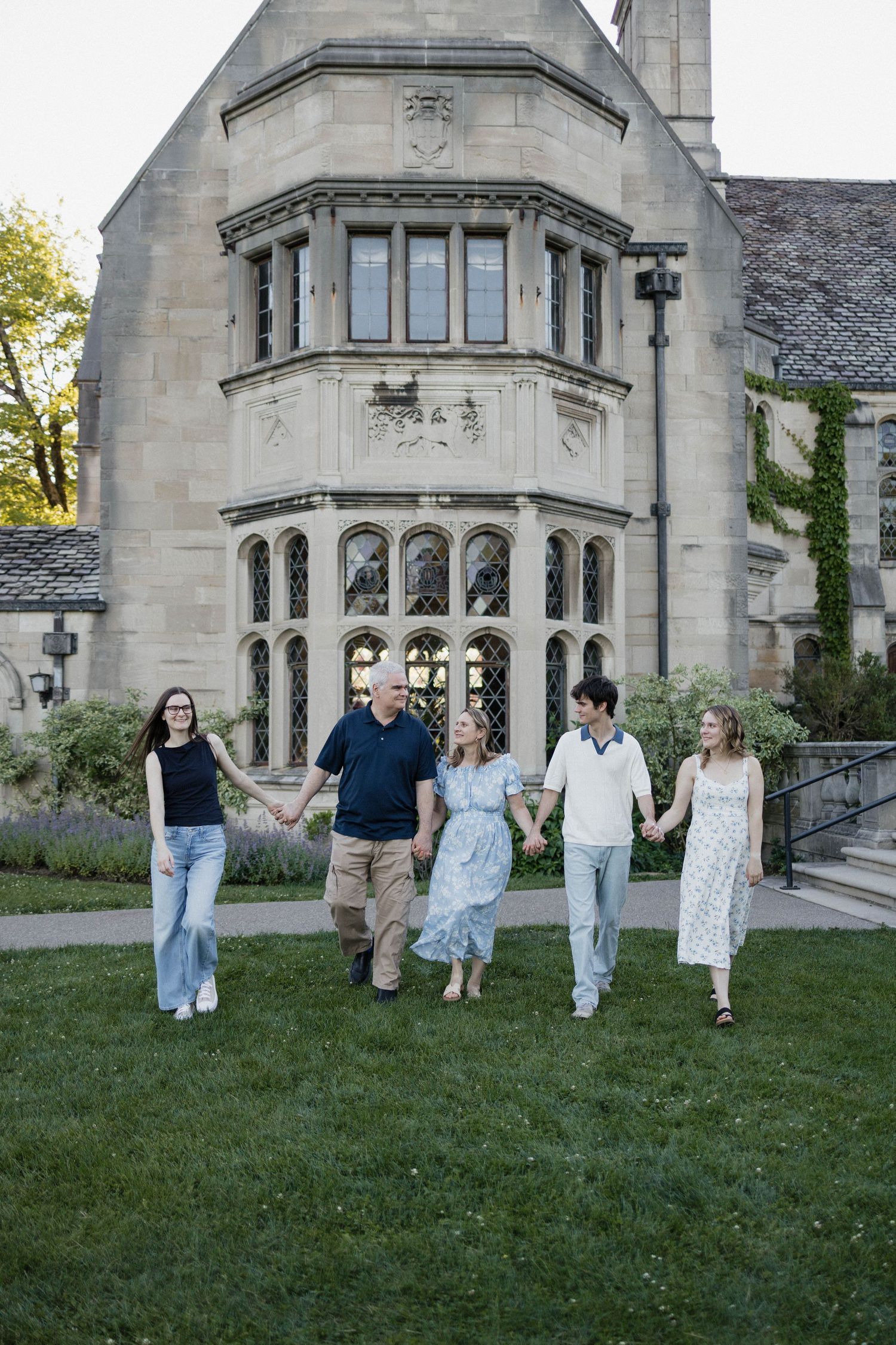 Family of five holding hands walking on grass in front of a grand stone building with Gothic architecture.