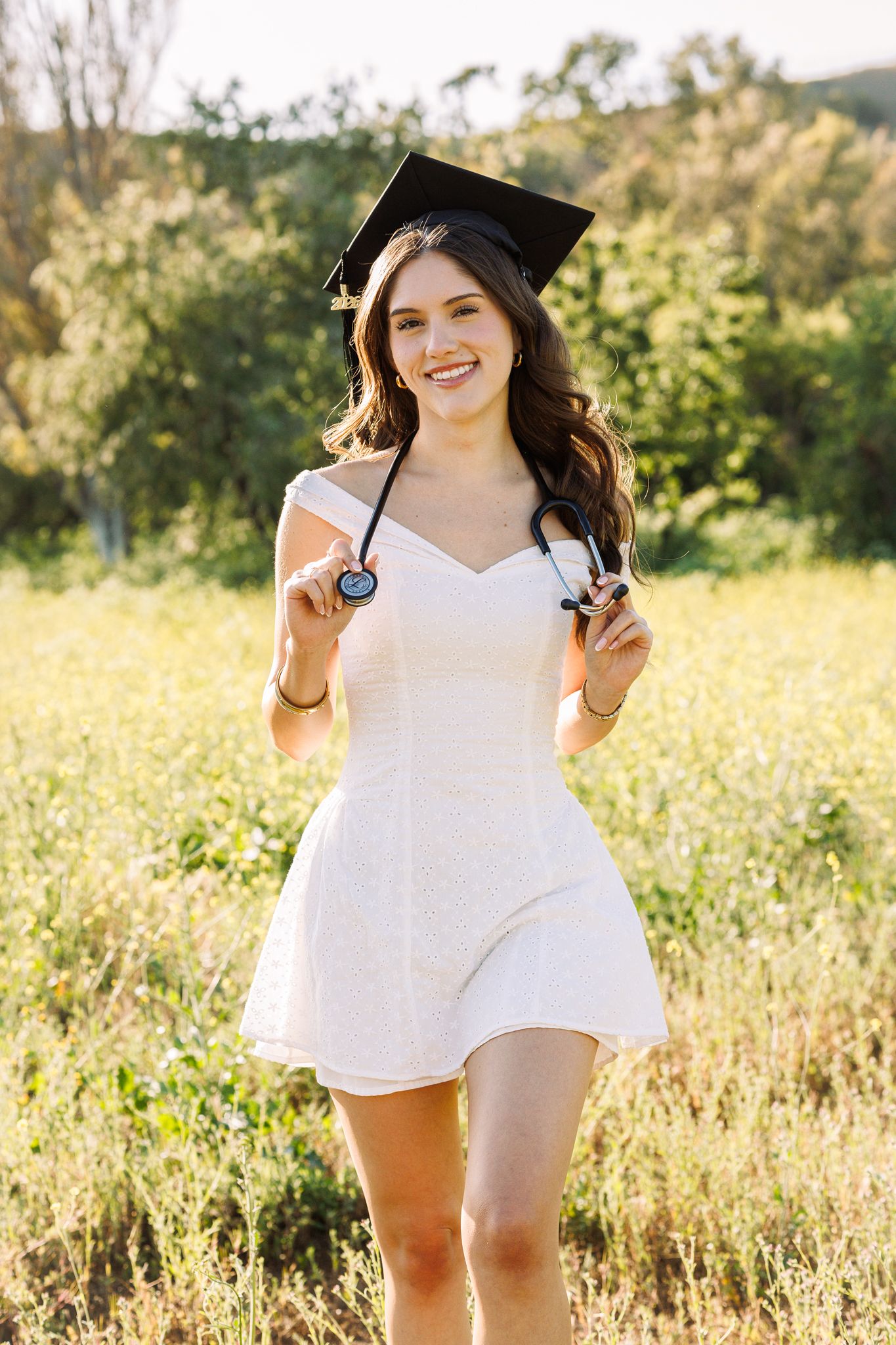 Smiling graduate in white dress and cap holds stethoscope outdoors in sunny field, celebrating medical school graduation.