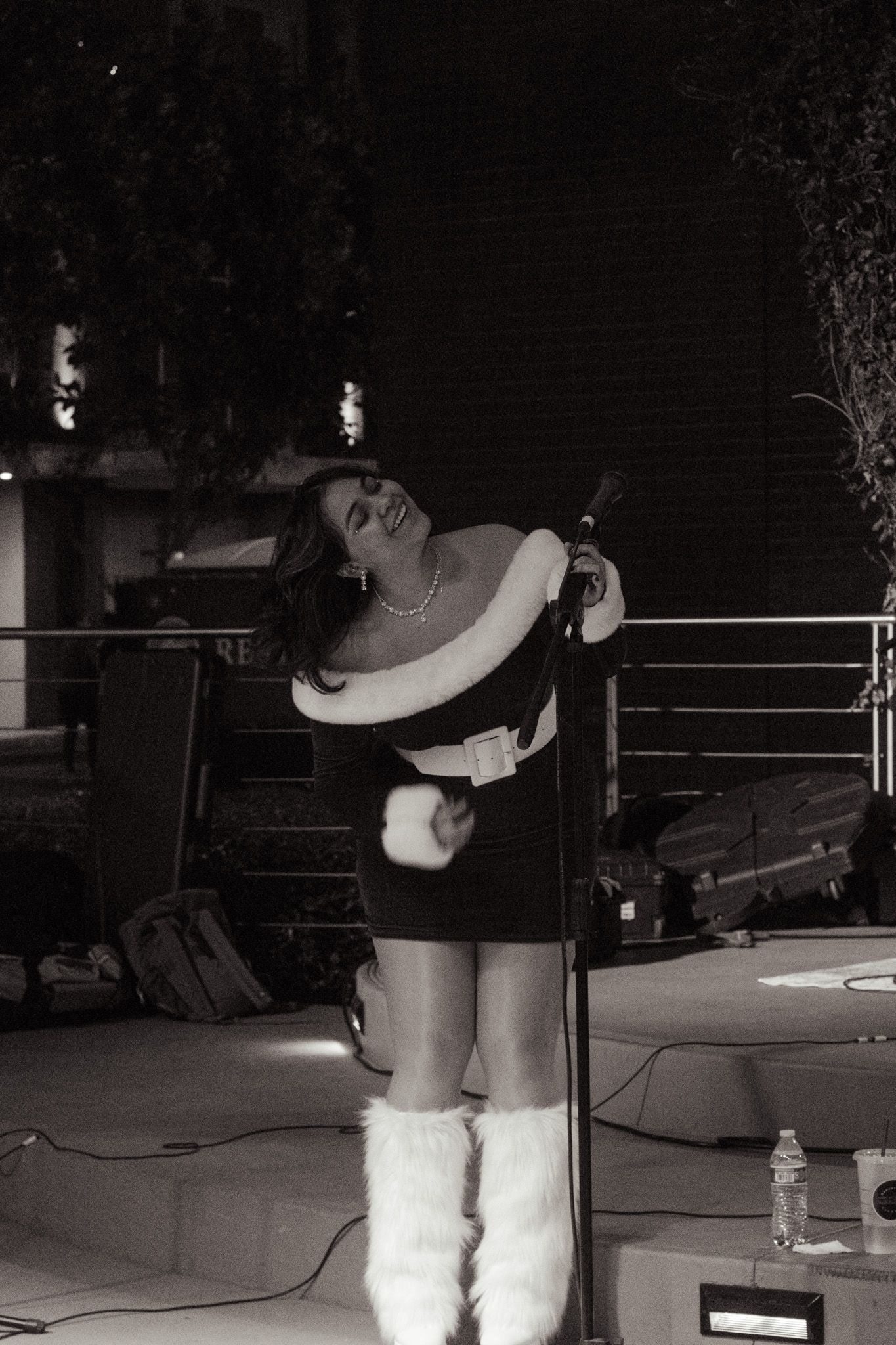 Performer in black dress and white furry boots sings passionately on stage at night with microphone and concert equipment.