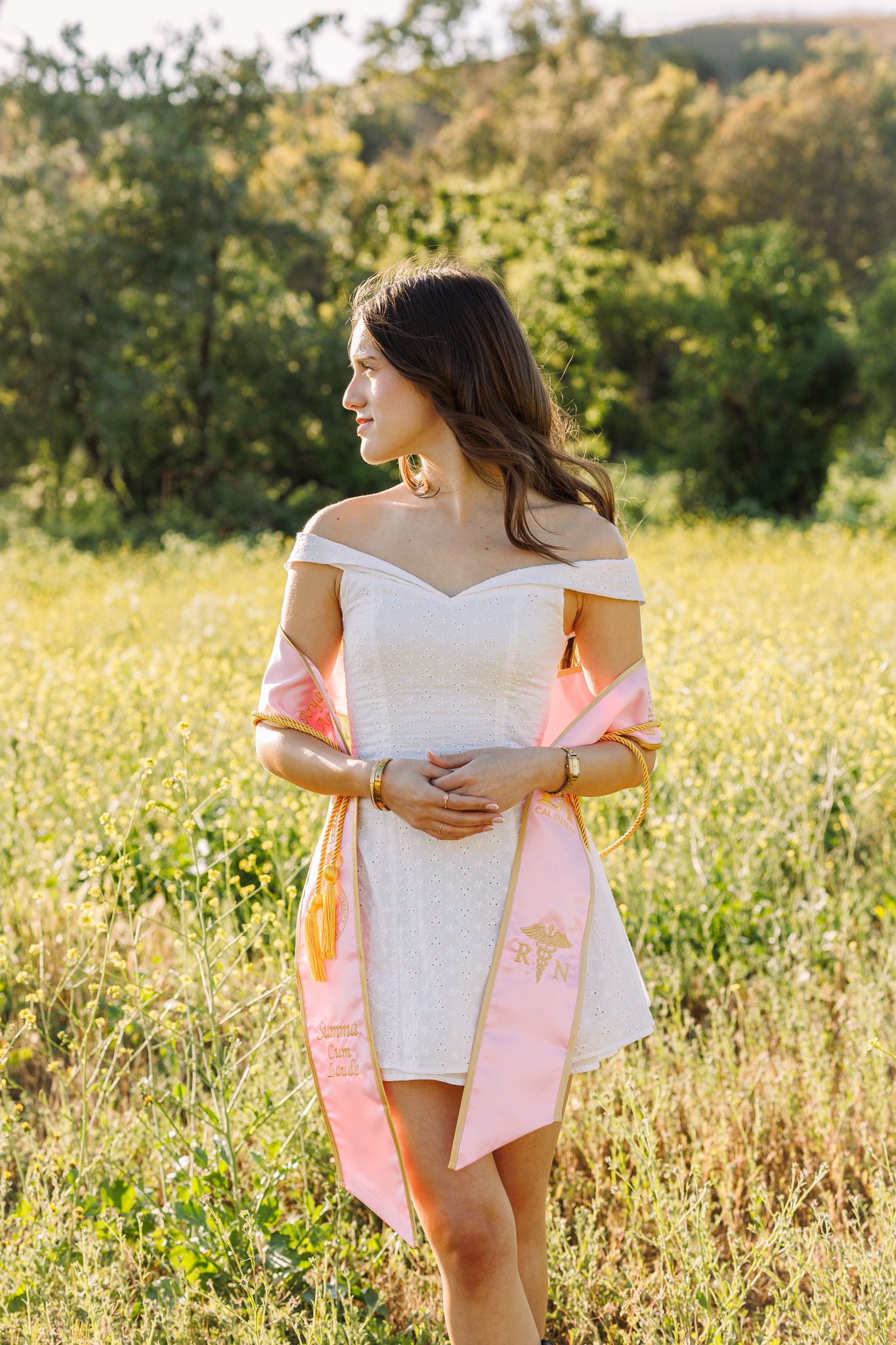 Young woman in white off-shoulder dress with pink shawl standing in a sunny green meadow field.
