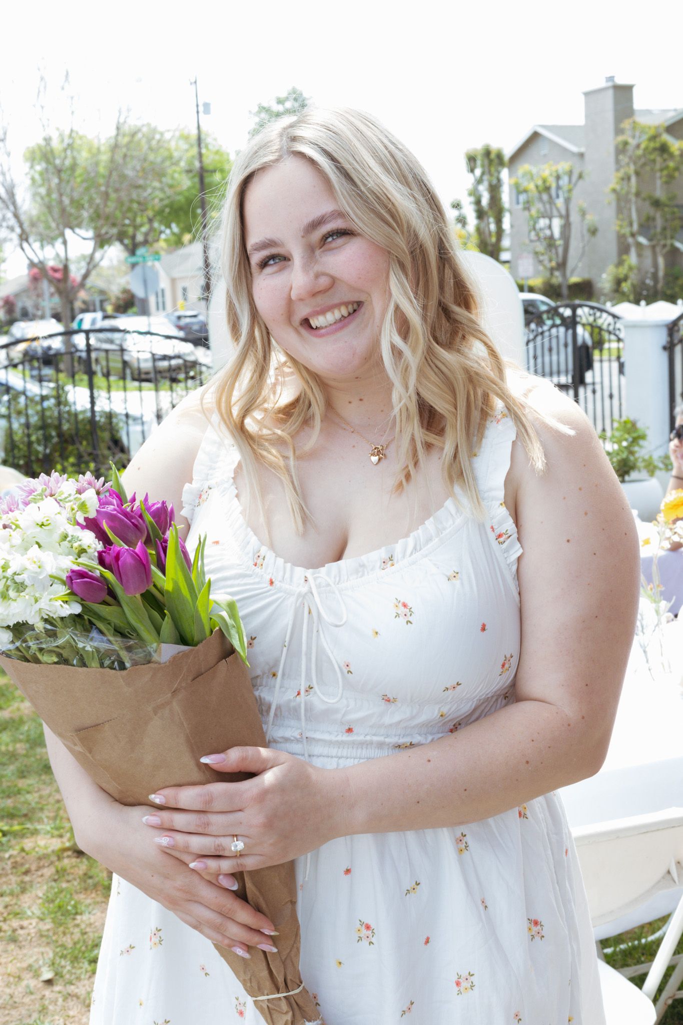 A bright spring scene shows someone in a white floral dress holding a bouquet of purple tulips outdoors.