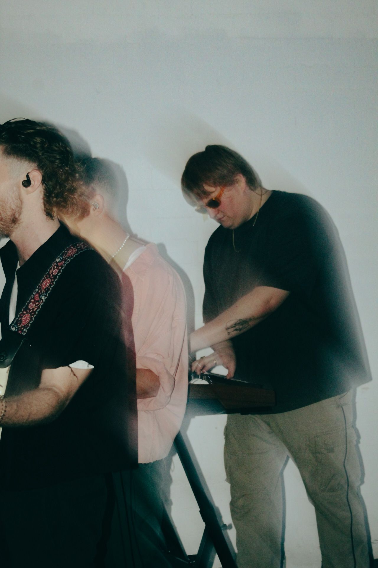 Three musicians in dimly lit studio working with instruments and equipment against white wall.