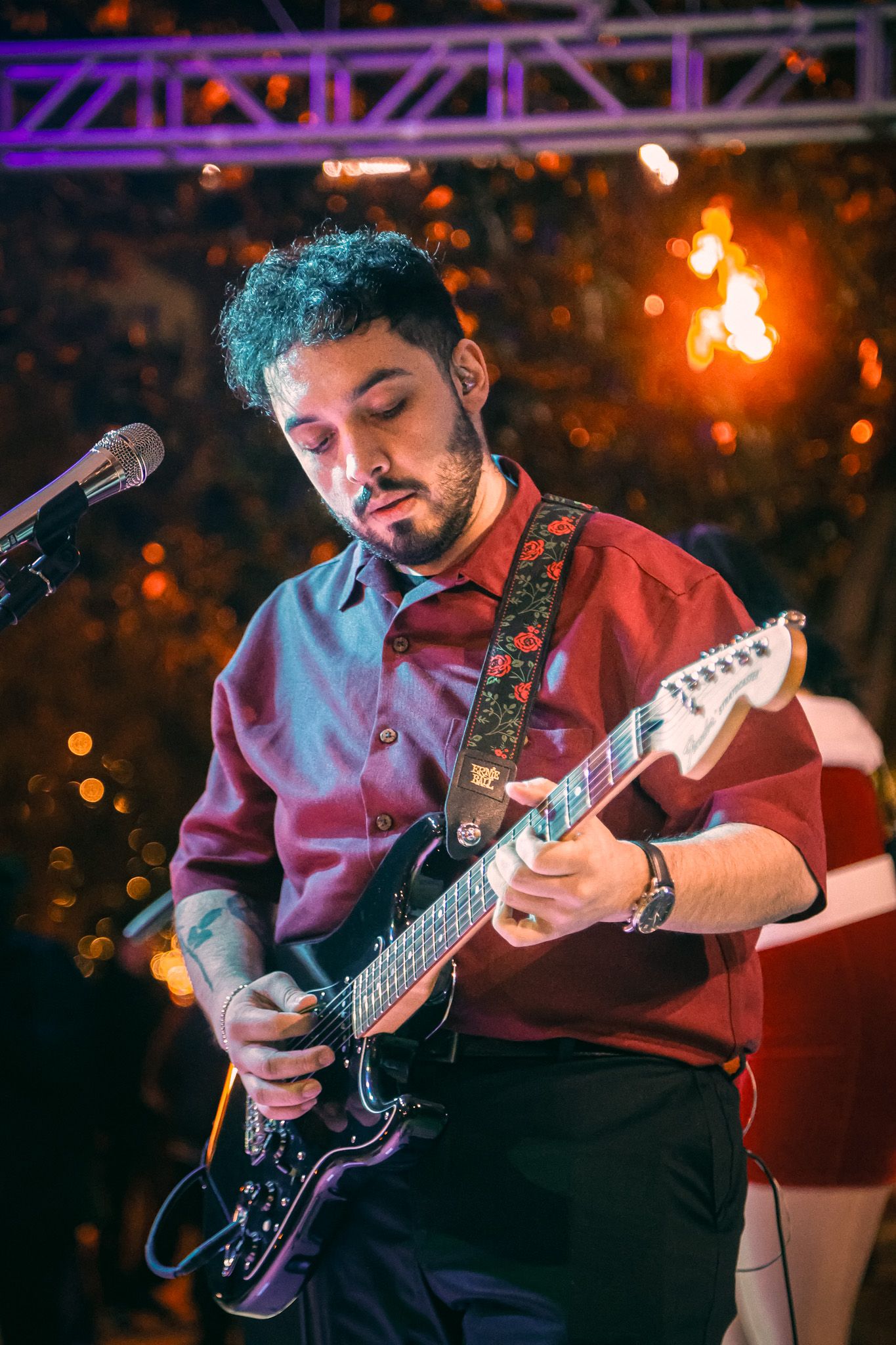 A musician in a red shirt plays electric guitar on stage with purple lighting and bokeh lights in the background.
