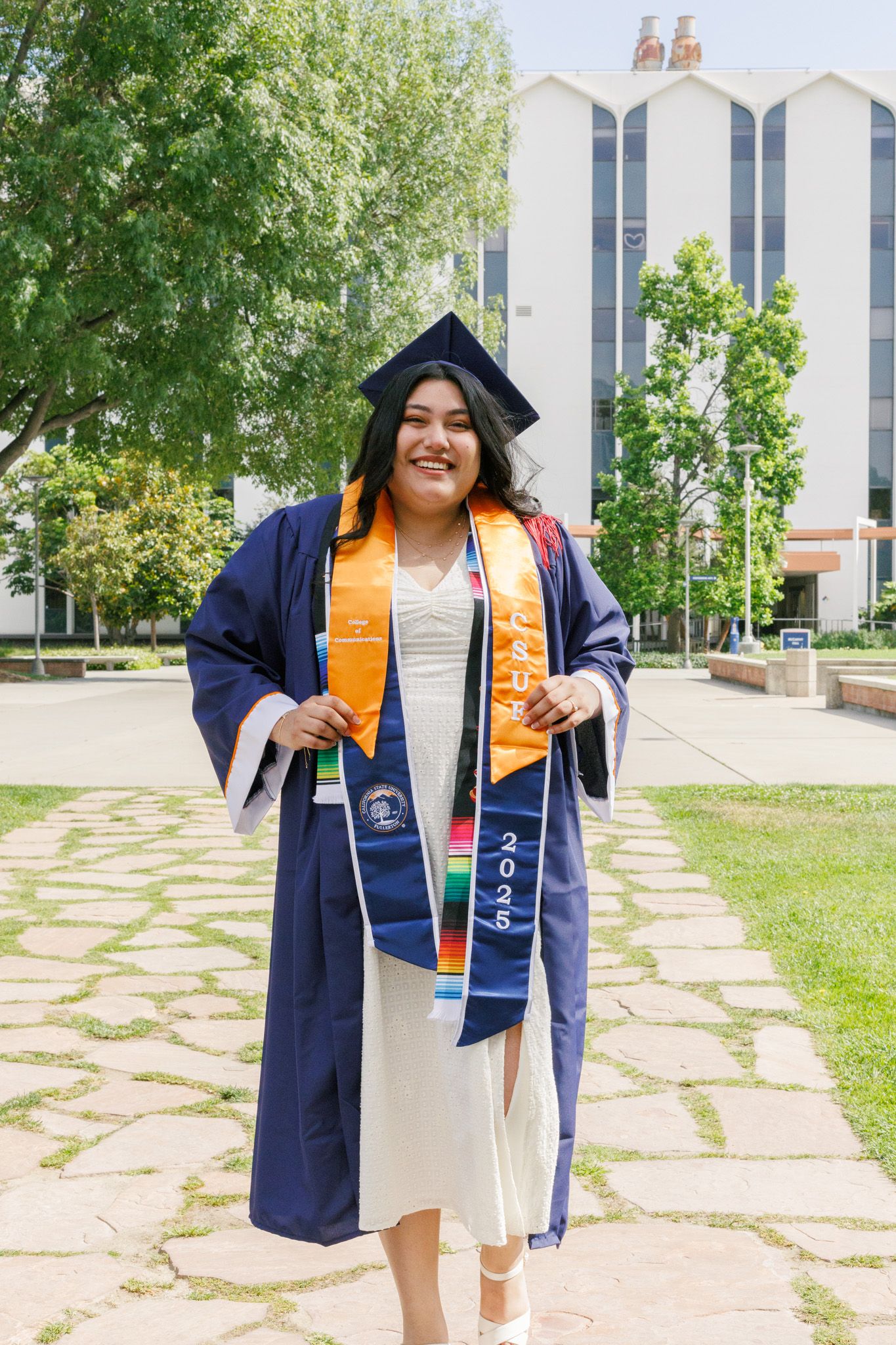 Graduate in navy cap and gown with gold honor stoles stands smiling on campus pathway near modern university building.