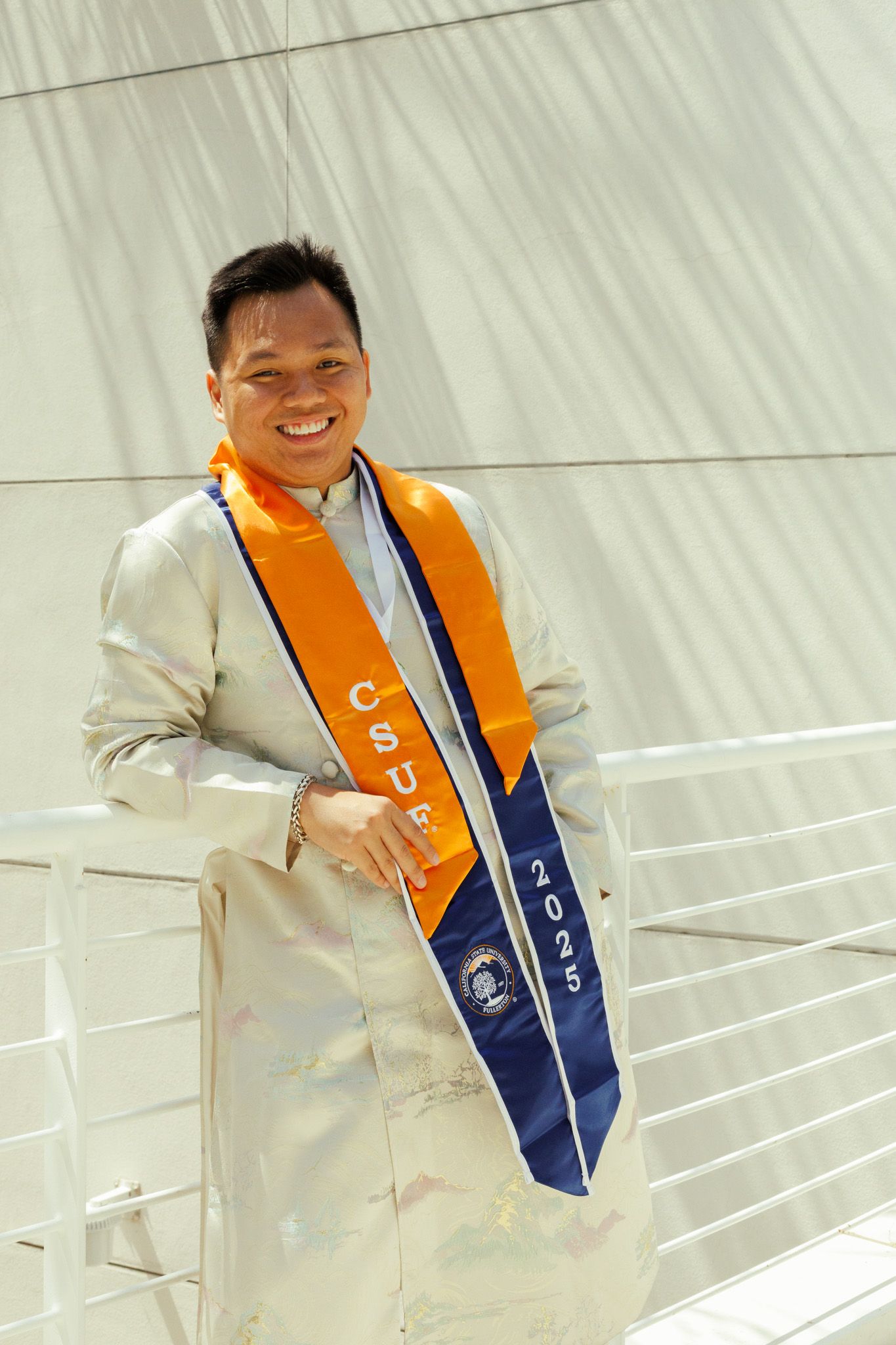 Graduate wearing a white suit and orange and blue graduation sash stands smiling against a light background.