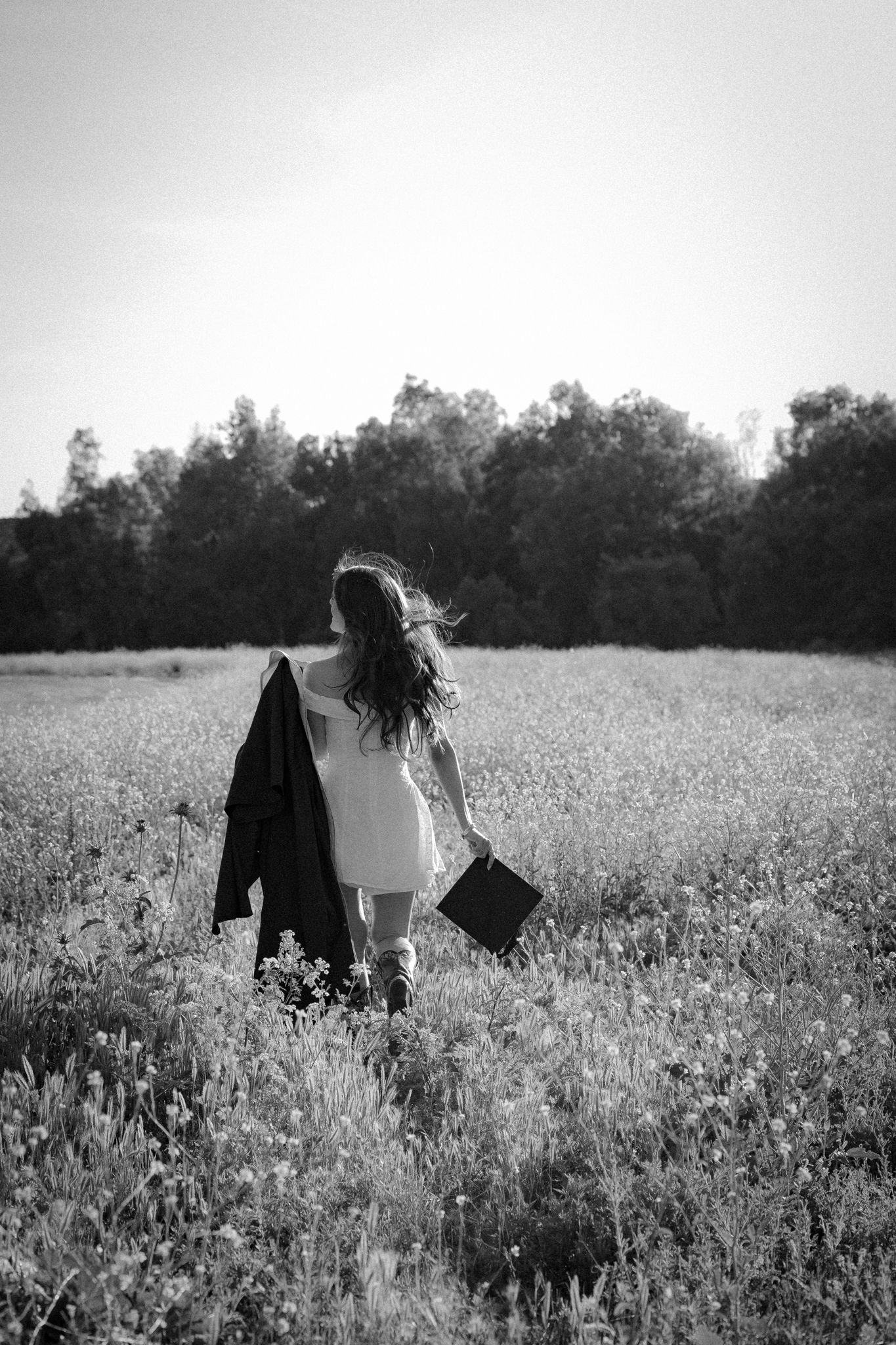 Black and white photo of a girl walking through a field carrying a suitcase and jacket toward distant trees.