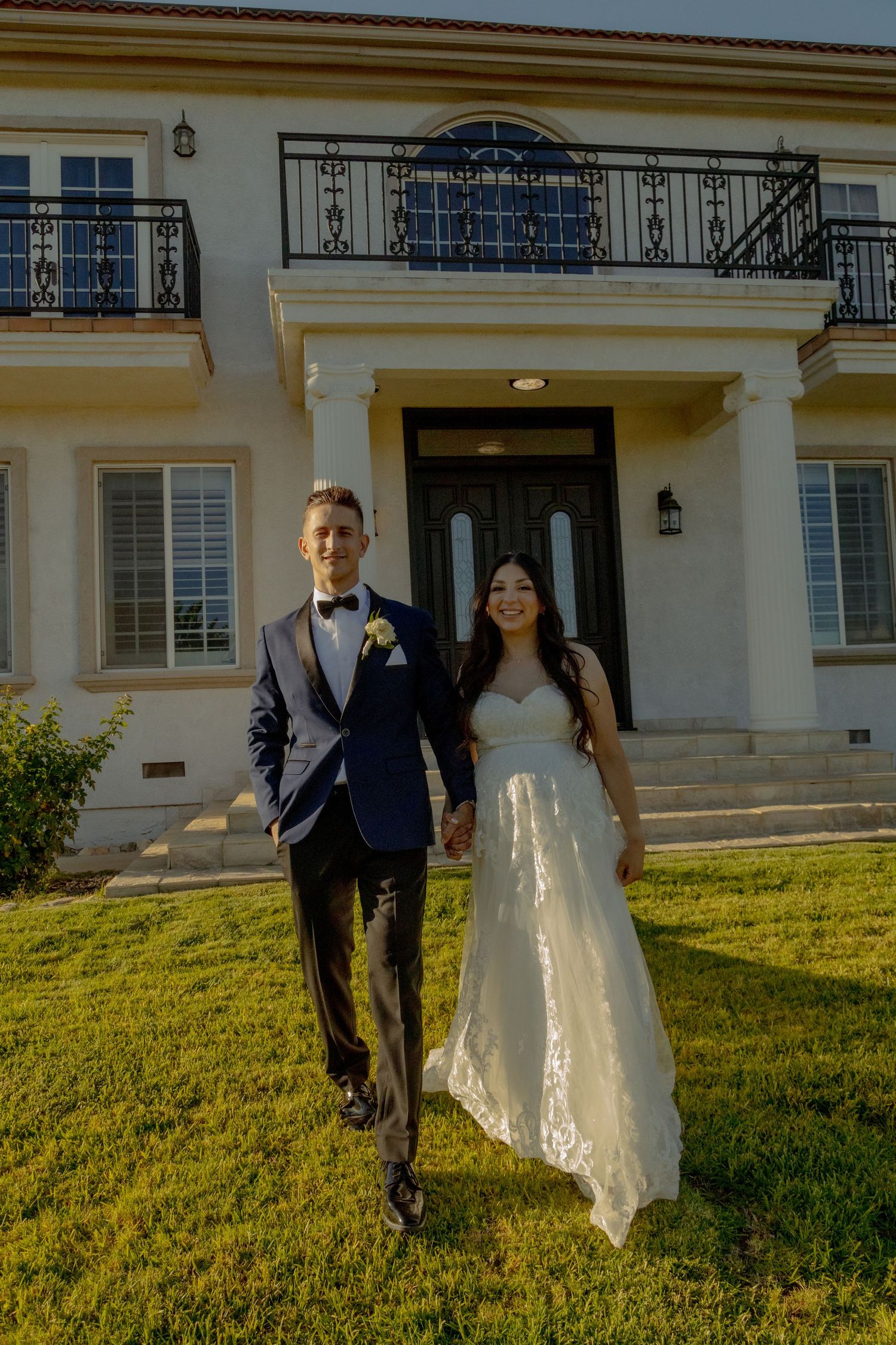 Newly married couple walks together on grassy lawn outside elegant mansion venue at golden hour.