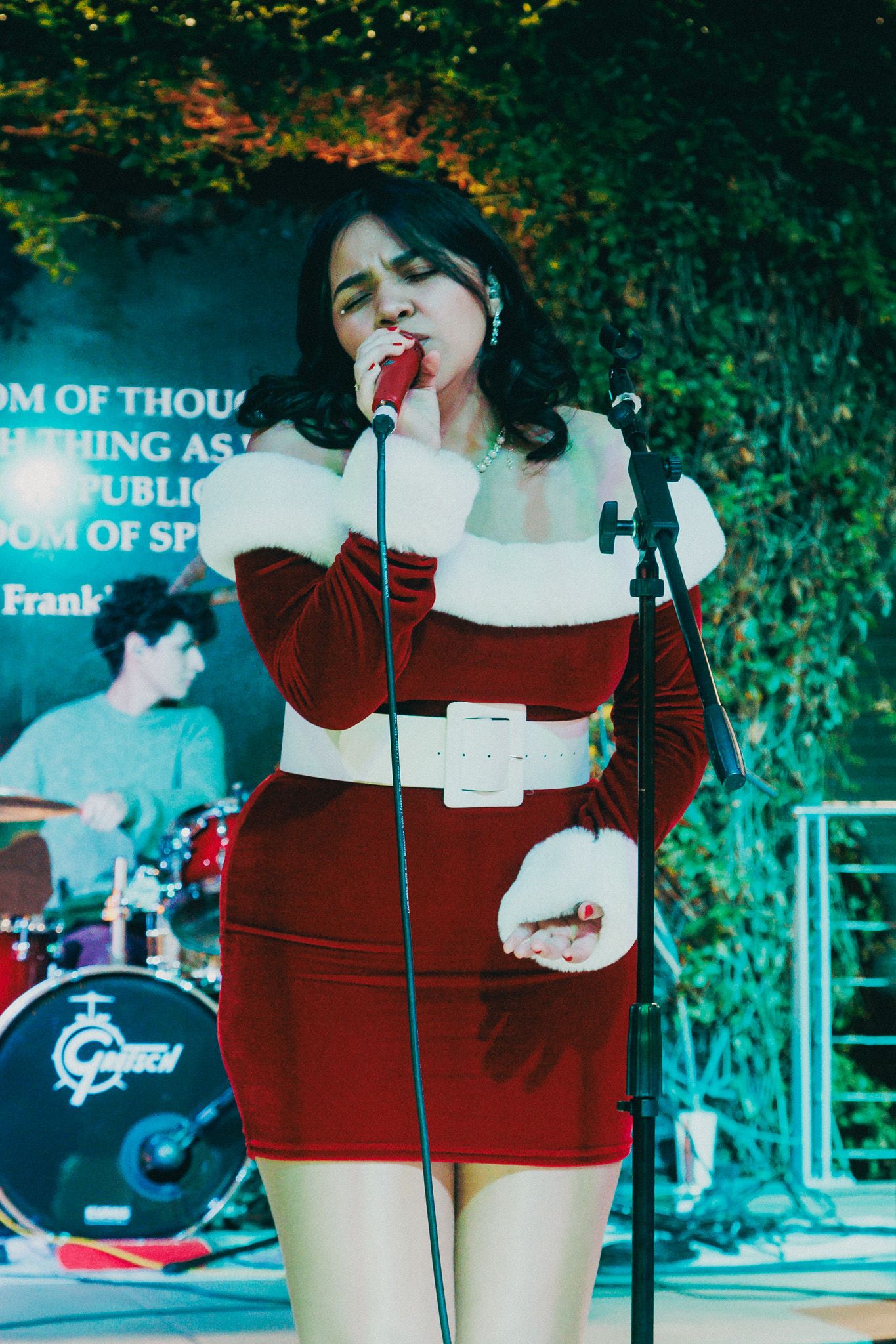 Singer performs on stage wearing a red and white dress with a microphone at an outdoor venue with greenery in background.