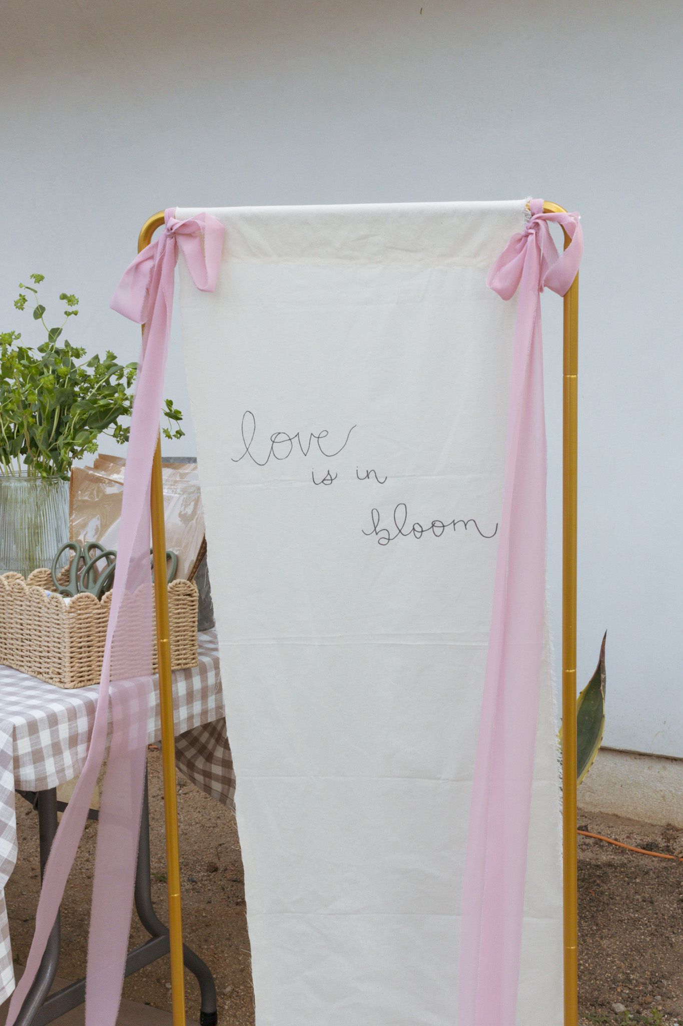 A white banner with 'love is in bloom' text decorated with pink ribbons stands next to potted plants and a checkered tablecloth.