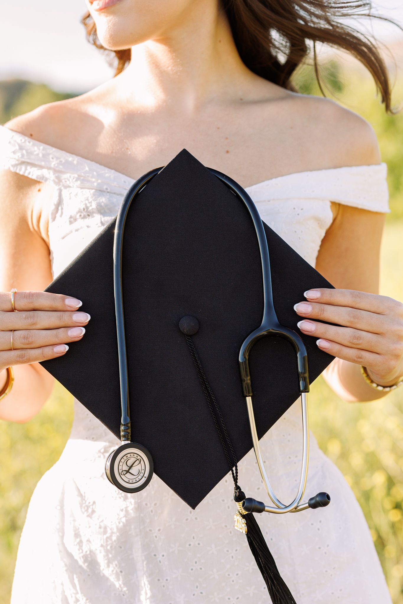 Woman in white dress holding black graduation cap with stethoscope, celebrating medical school graduation outdoors.