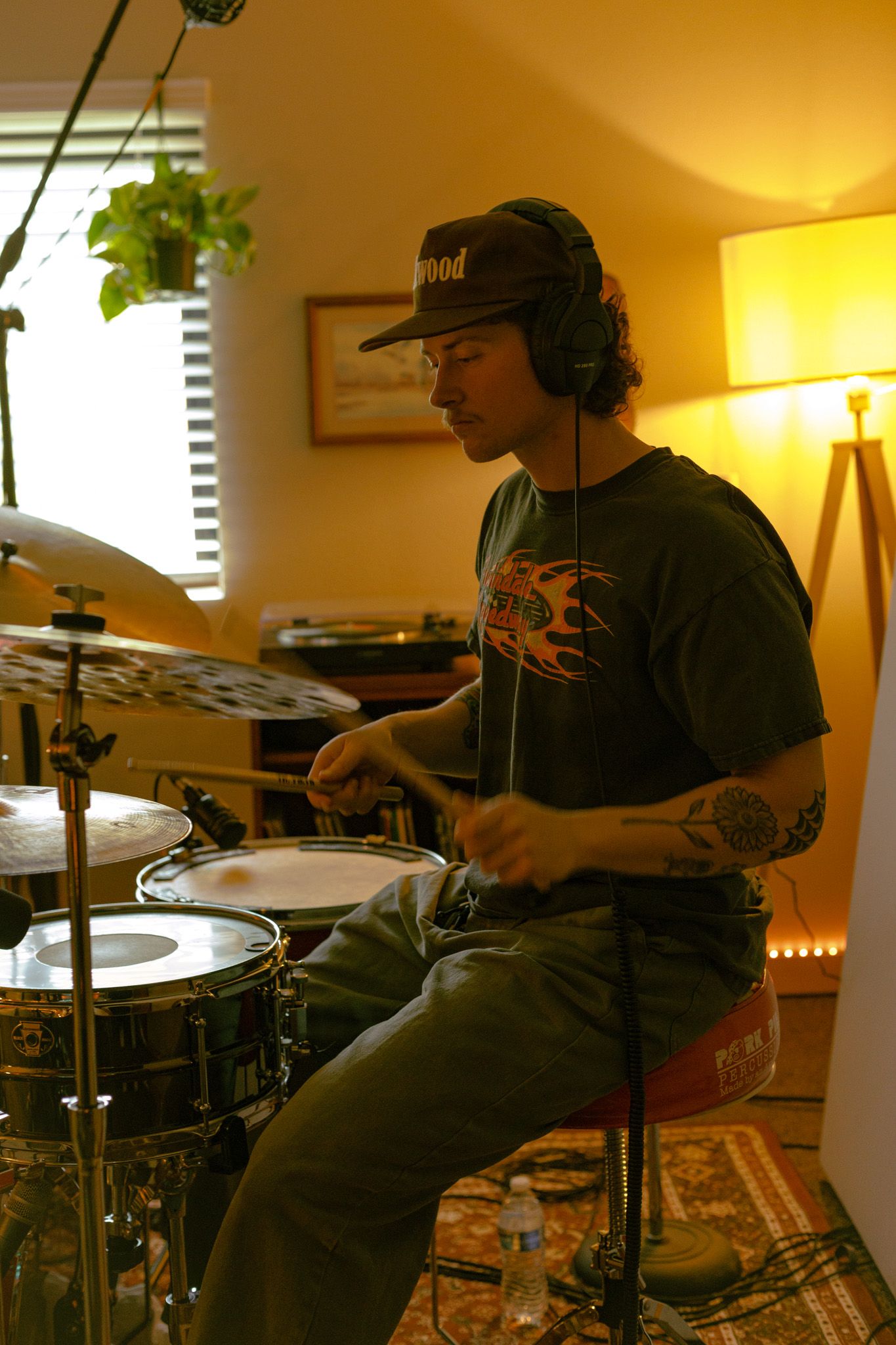 A drummer wearing headphones and a baseball cap plays an electronic drum kit in a warmly lit room with a houseplant nearby.