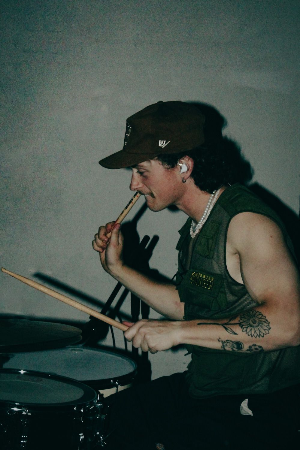 A drummer in a dark baseball cap and tank top plays while holding a drumstick in their mouth during a performance.