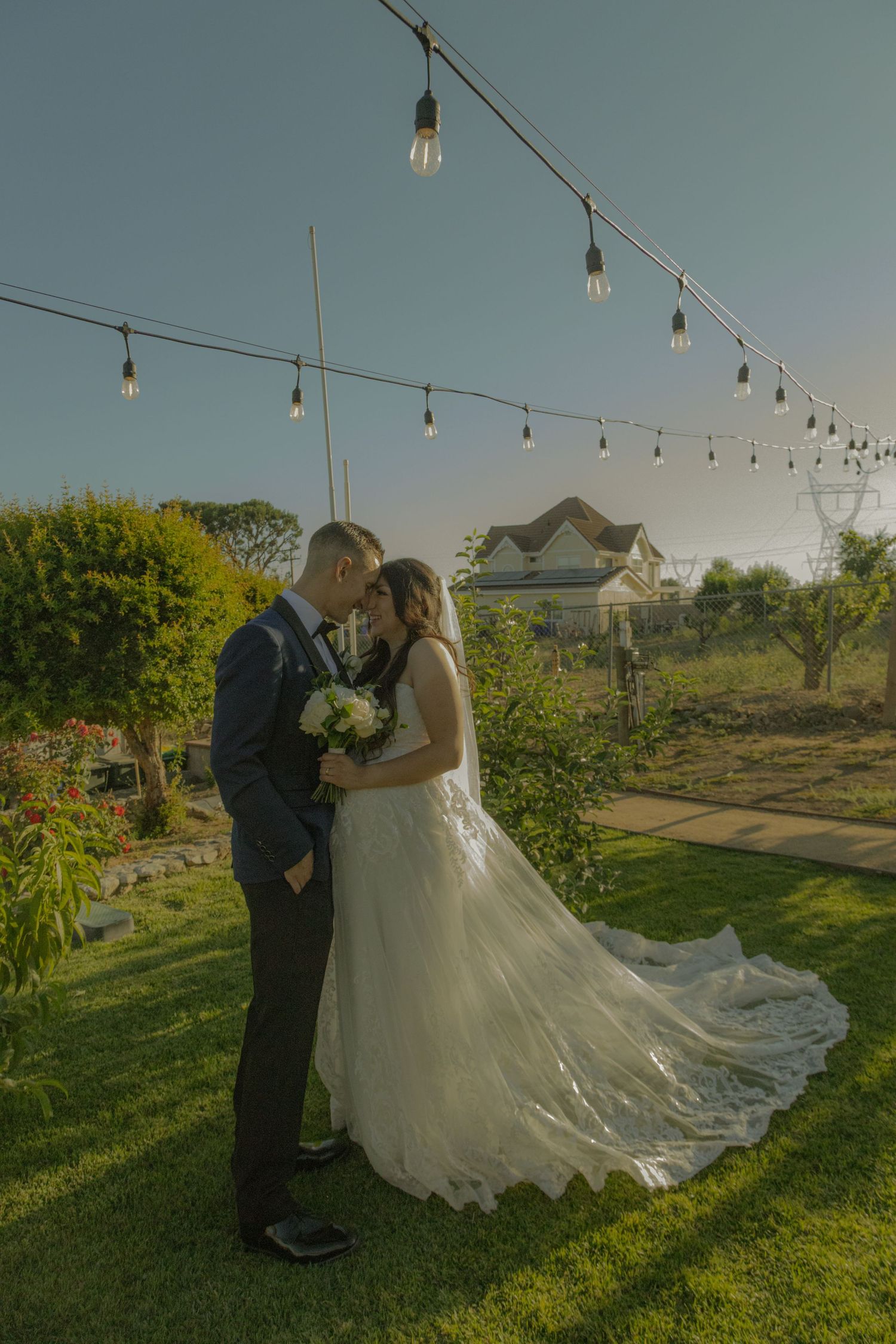 A romantic outdoor wedding scene on a lawn with string lights overhead and a flowing white gown at sunset.