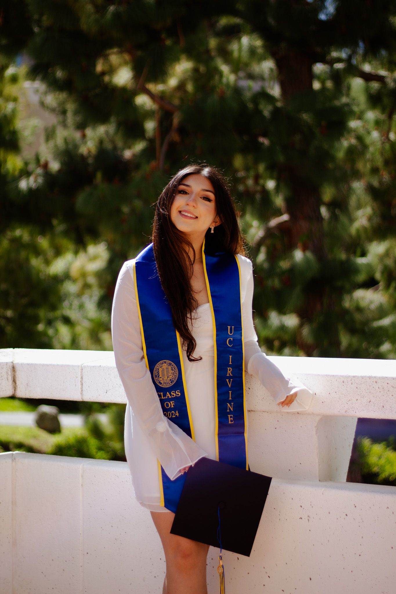 Graduate wearing white dress and blue graduation stole holds mortarboard while posing outdoors on sunny day.