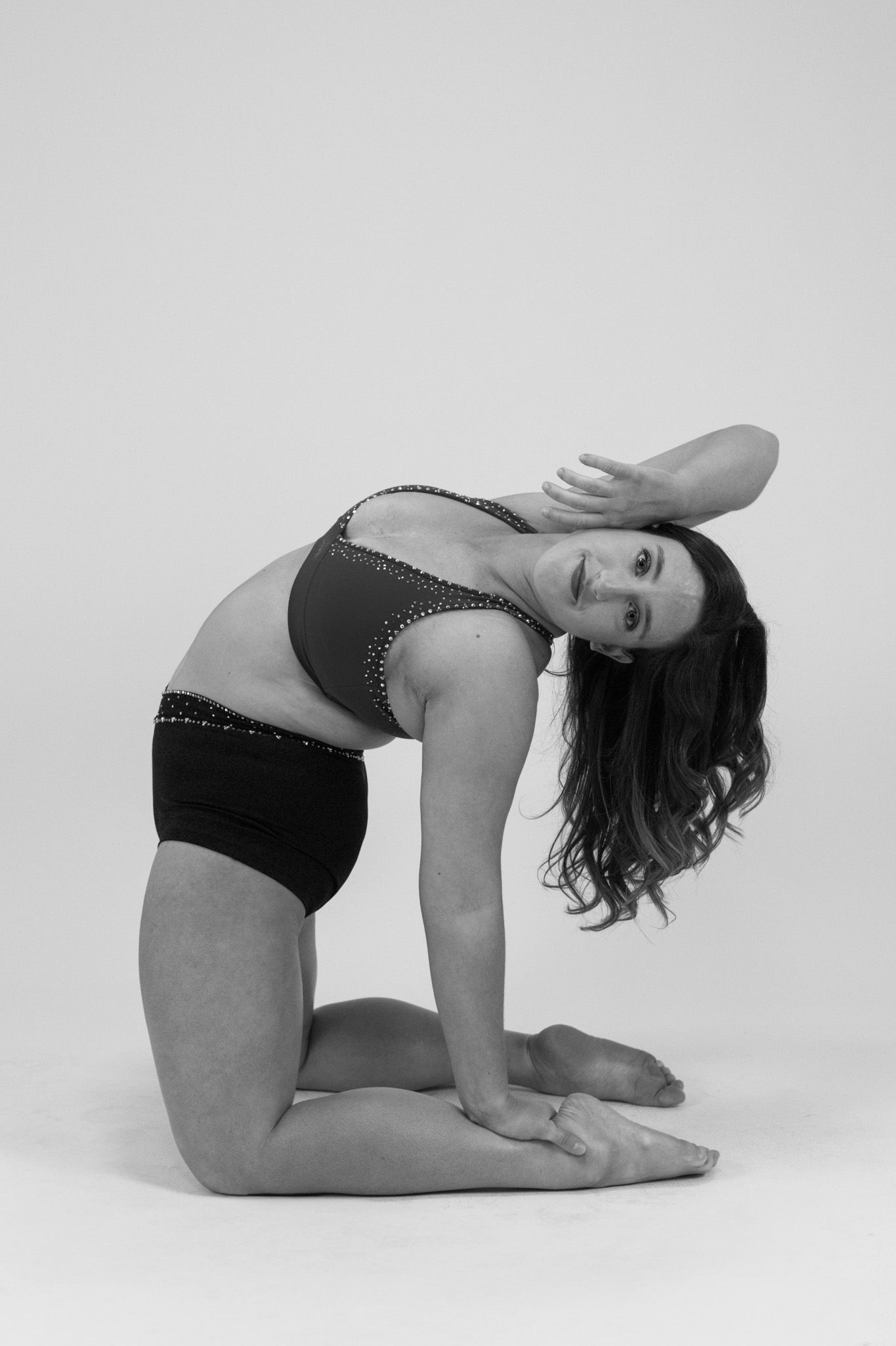 Black and white portrait photo of a dancer in athletic wear performing a cat pose yoga position against a plain backdrop.